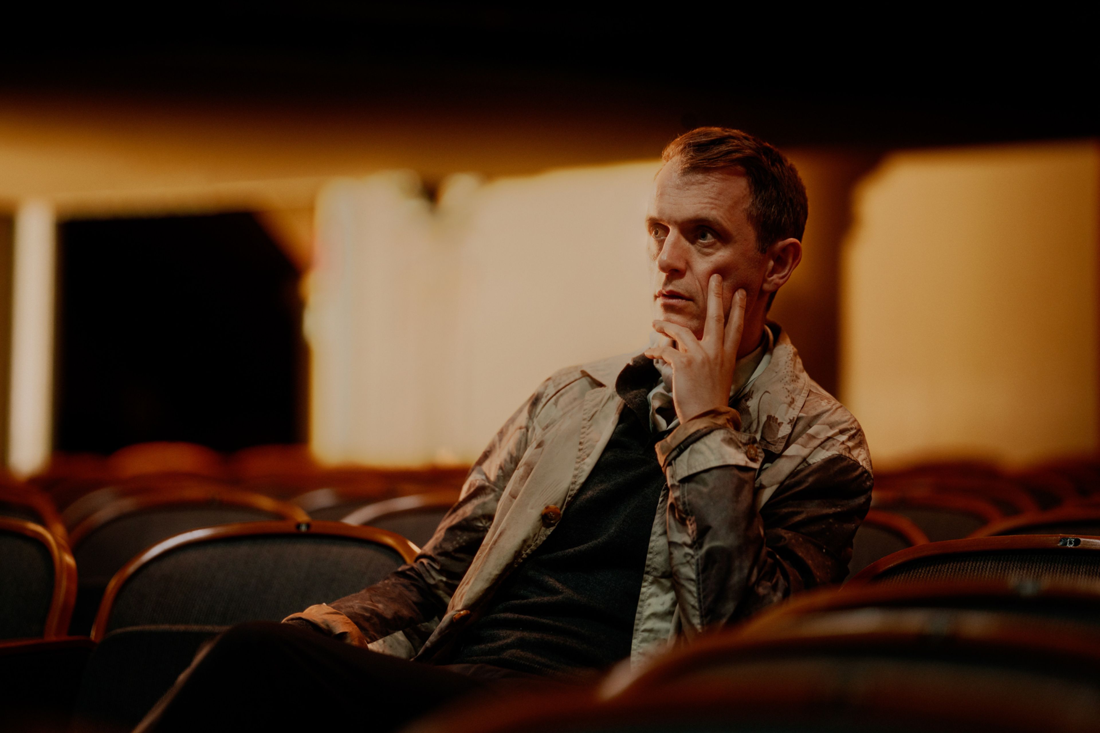 Nils Bech sitter i salen på Centralteatret og tenker med to fingre hvilende mot kinnbeinet. Belysningen er mørk og dus oransj