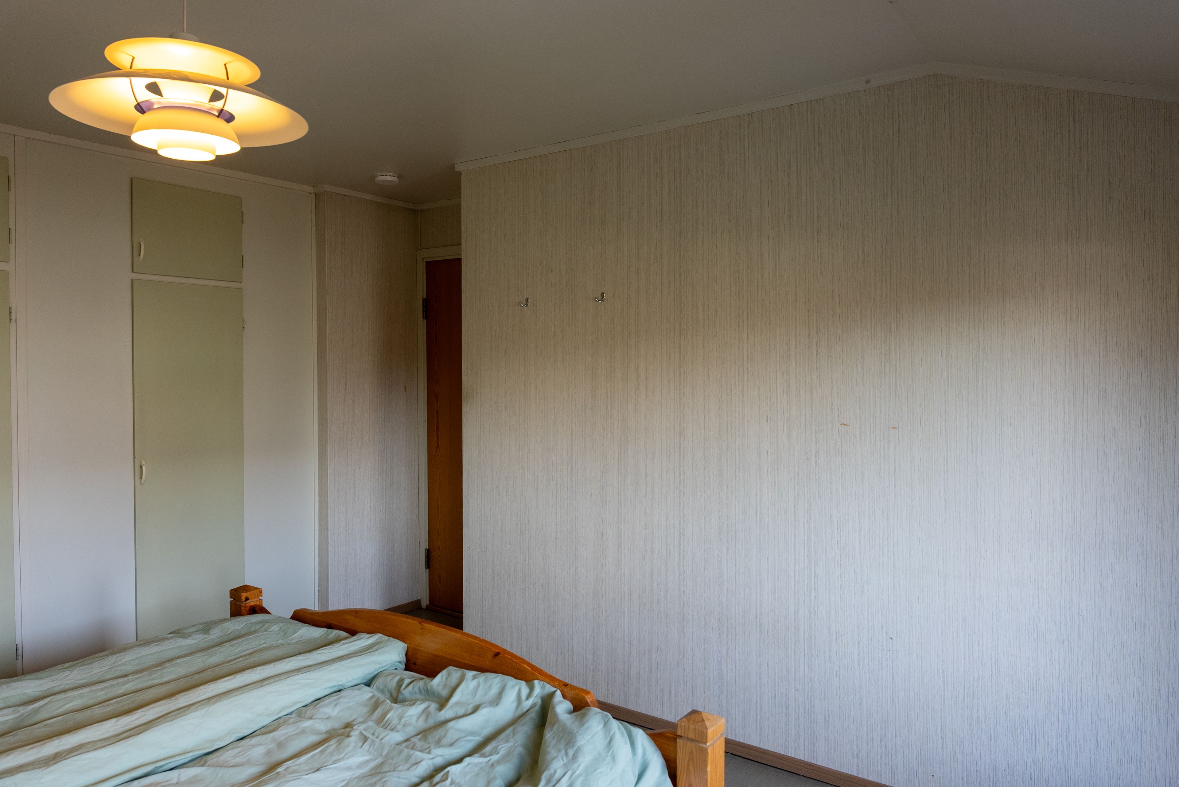 Eldre soverom med blomstrete tapet, linoleumsgulv, klesskap og furuseng.