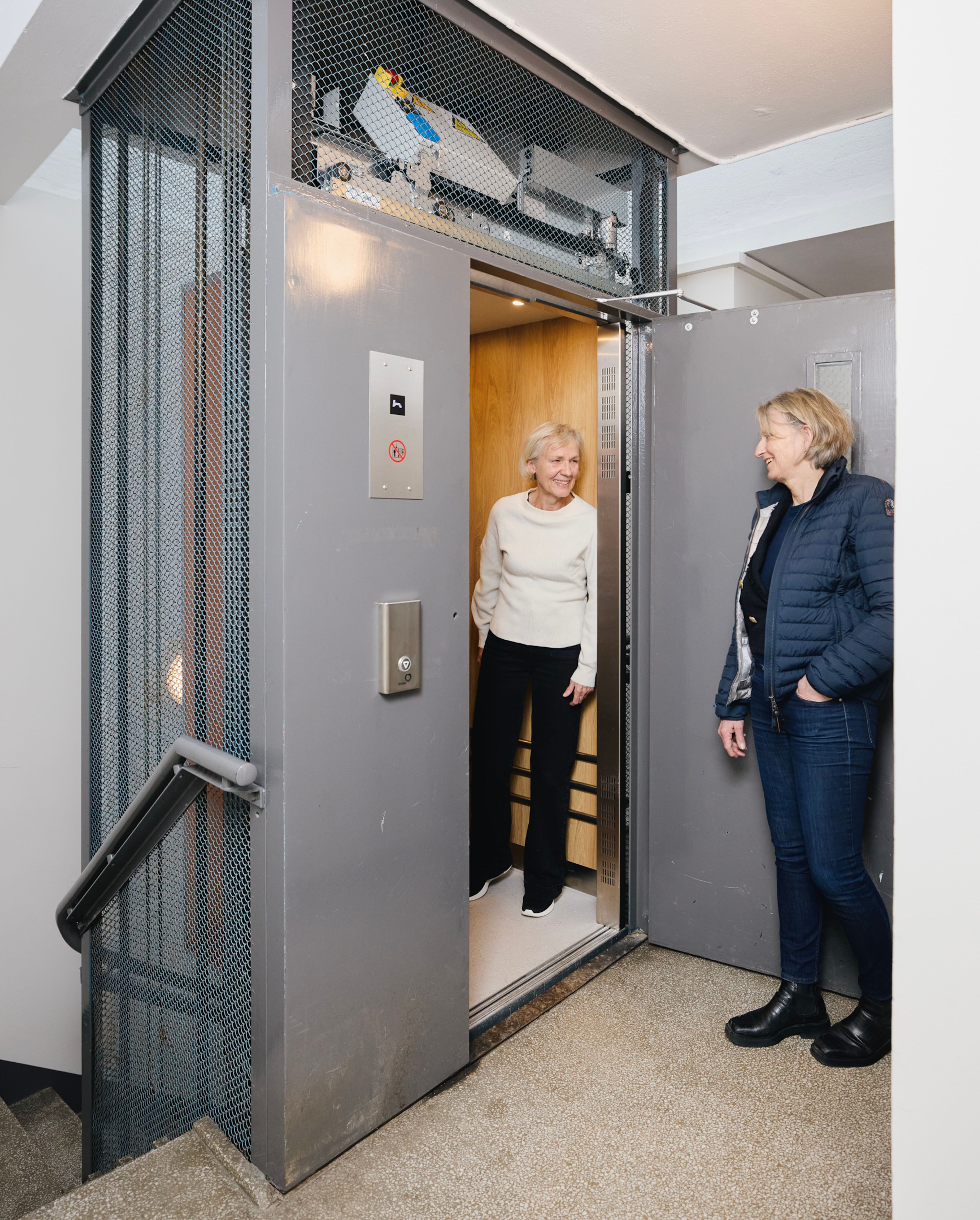 To kvinner står ved inngangen til en heis i en oppgang i et borettslag