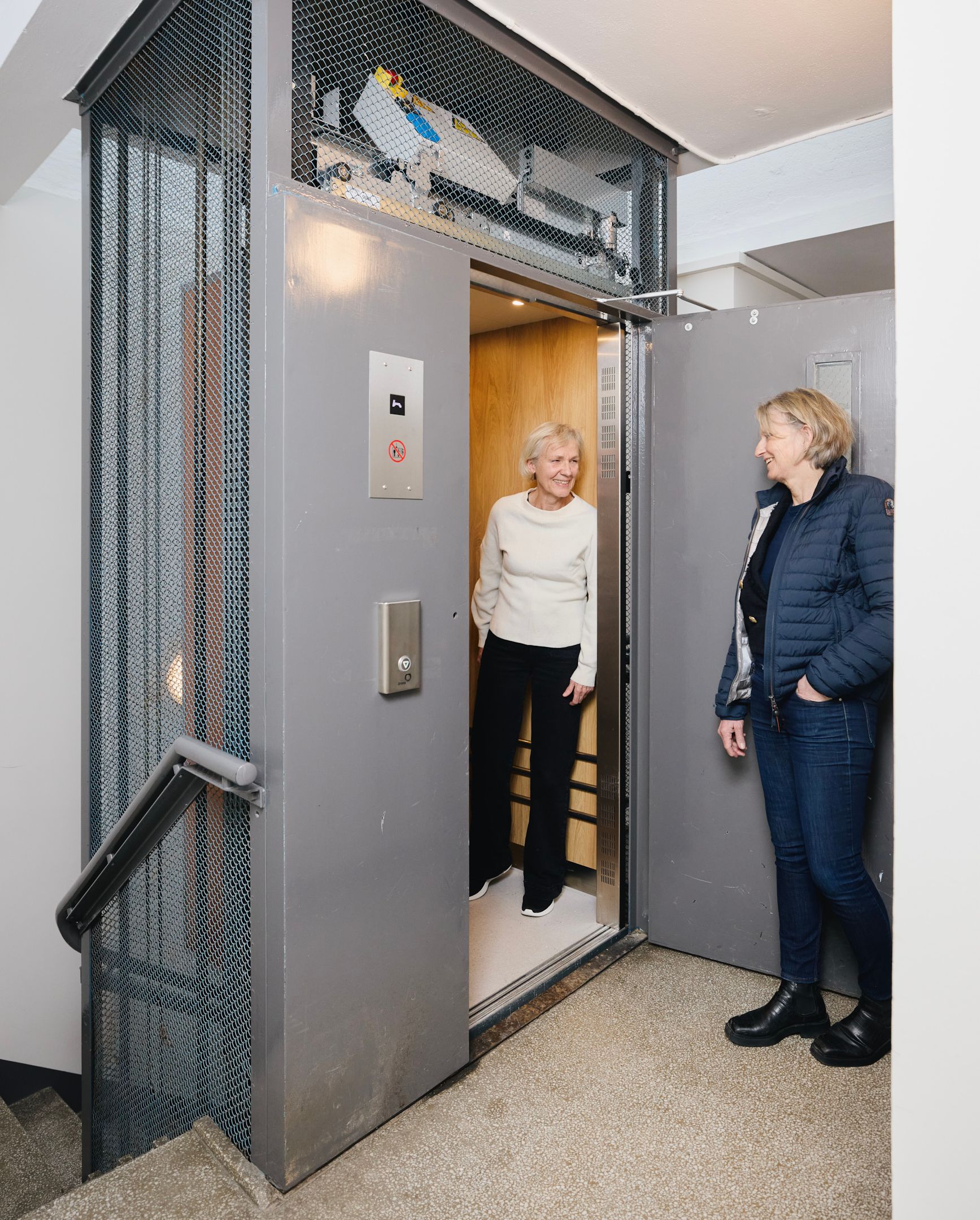 To kvinner står ved inngangen til en heis i en oppgang i et borettslag