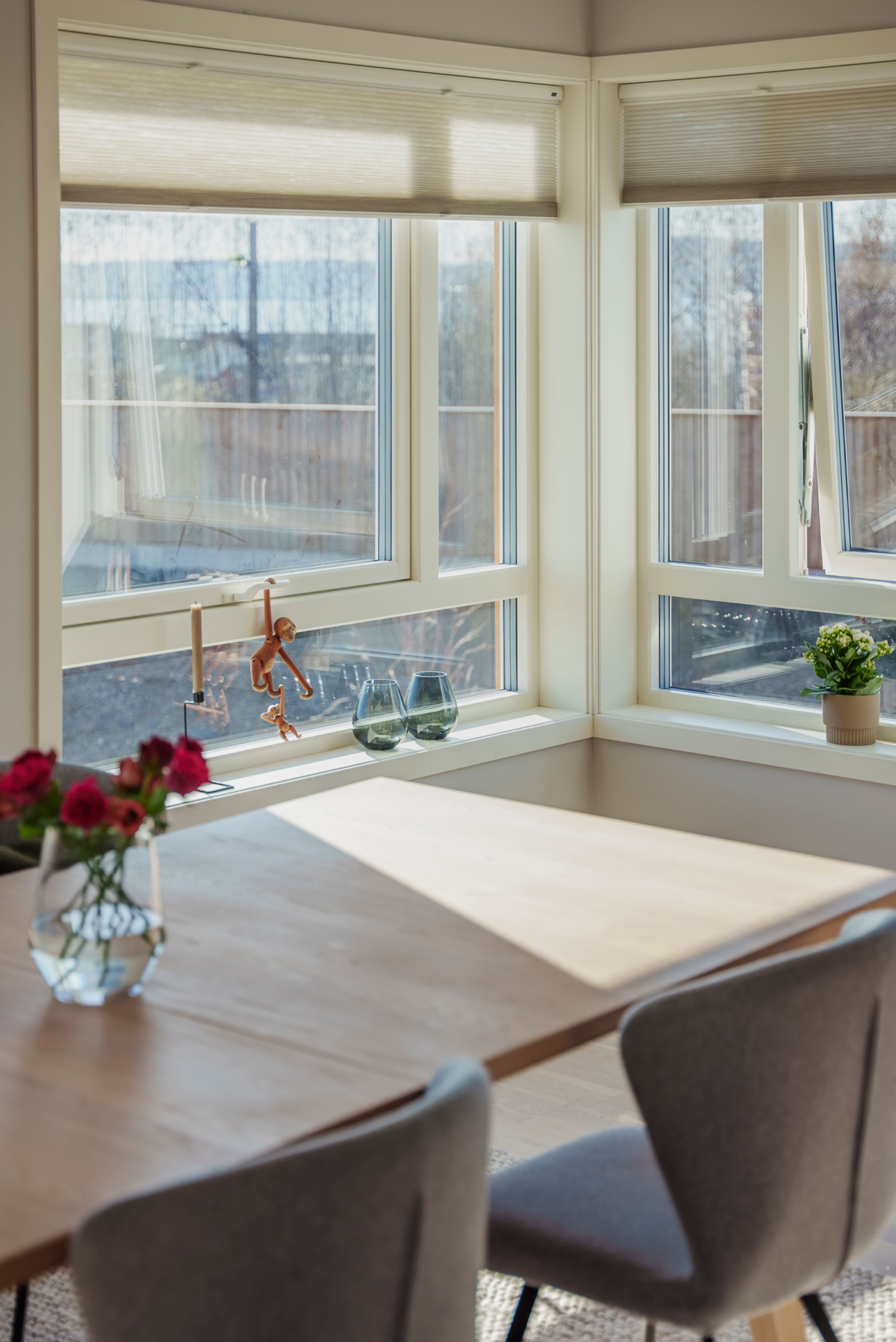 Lysebrunt spisebord foran en vindusrekke som slipper inn mye sol. På bordet er det en vase med røde roser.