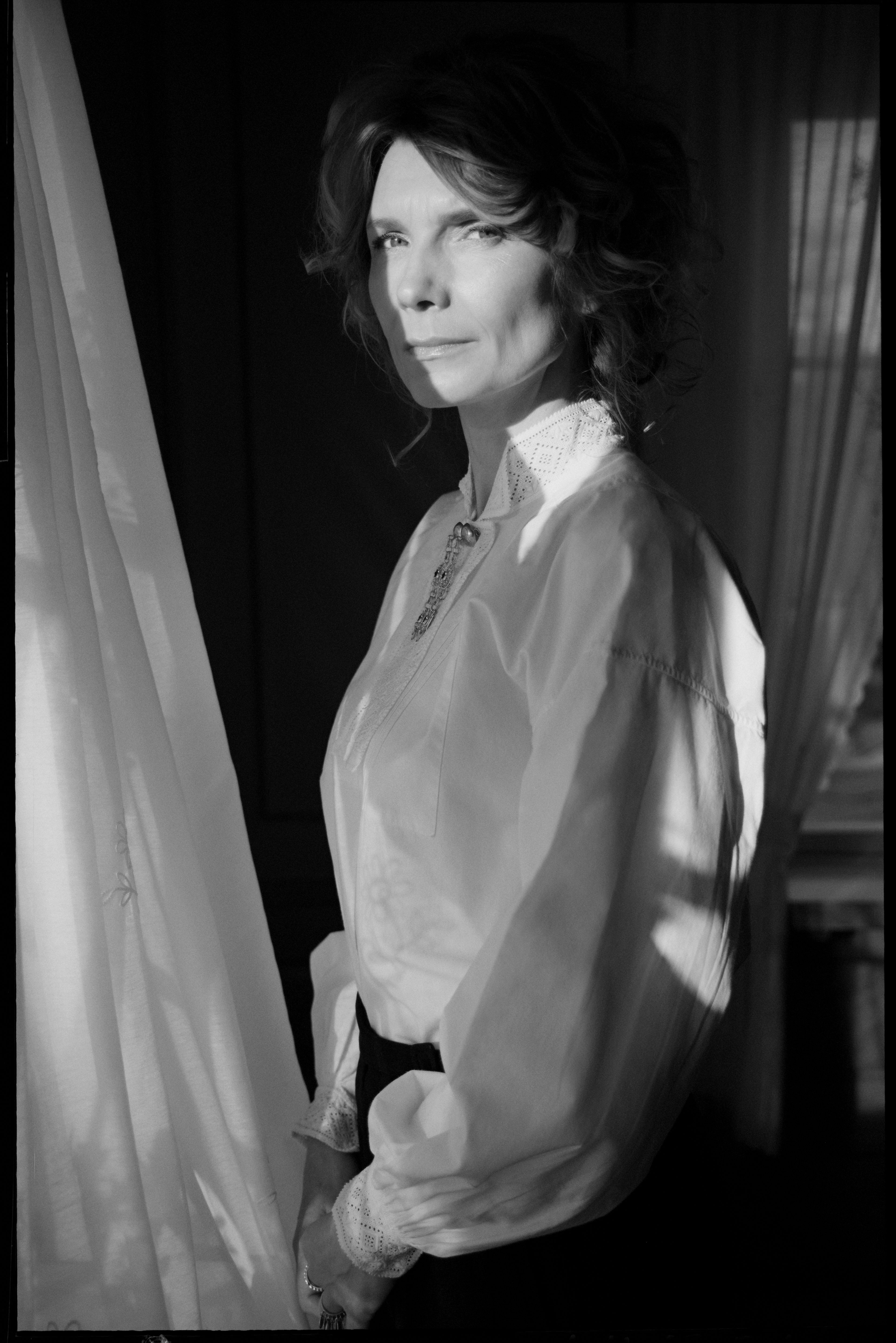 Et svart/hvitt bilde av Herborg Kråkvik. Hun står inntil en gardin og har på seg bunadskjorte.