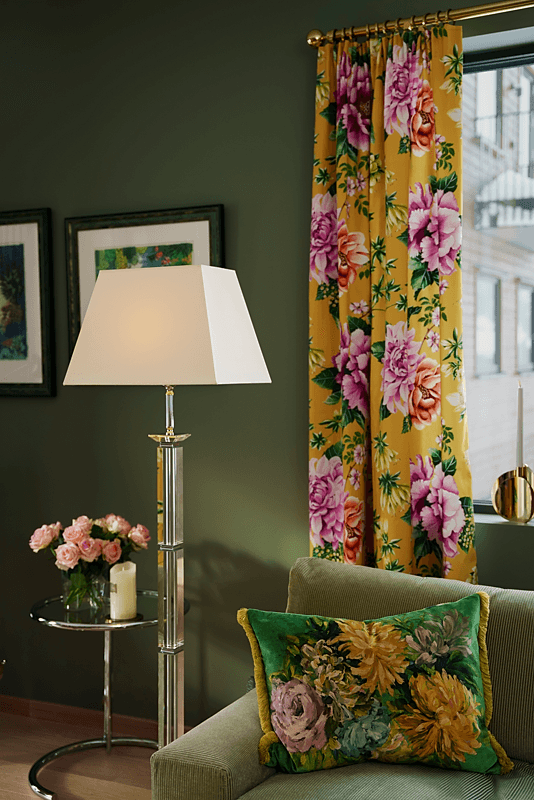 Detaljbilde av den ene stuelampa mot den grønne veggen og sofaen, til høyre for noen gulblomstrede gardiner