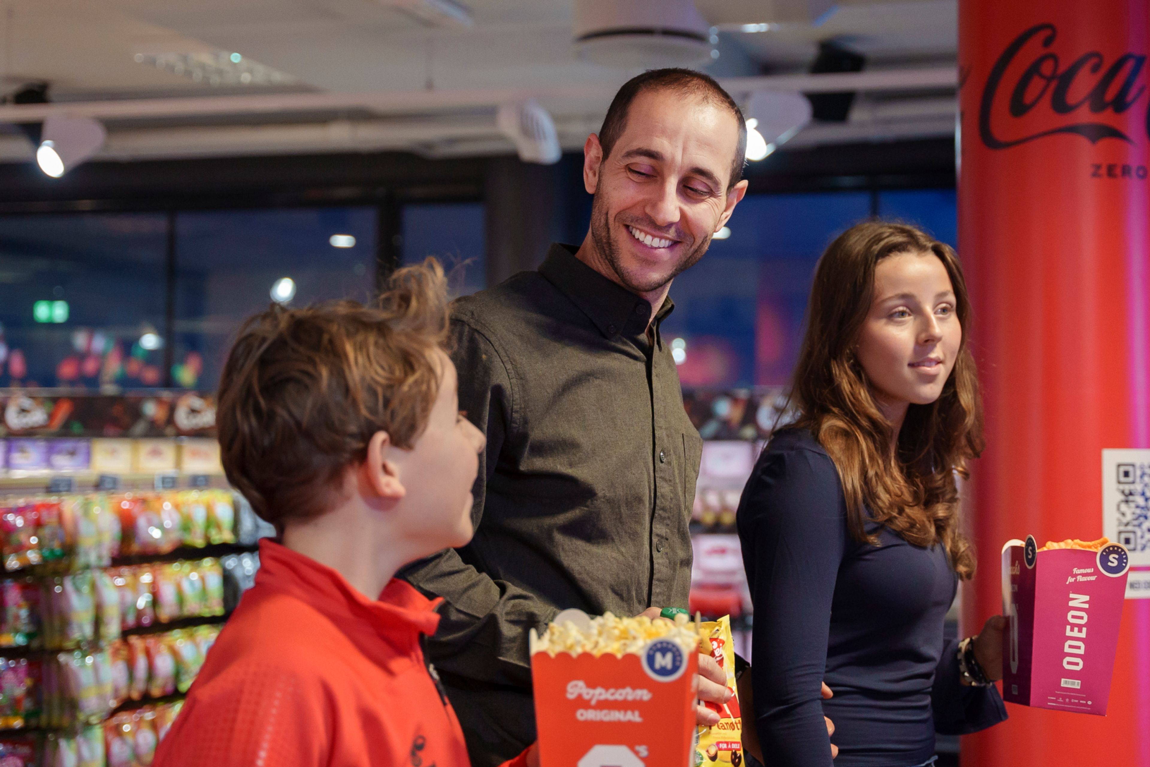 En mann er på kino med barna sine. En gutt og en jente holder i hver sin popcorn. Alle tre smiler og ser forventningsfulle ut.