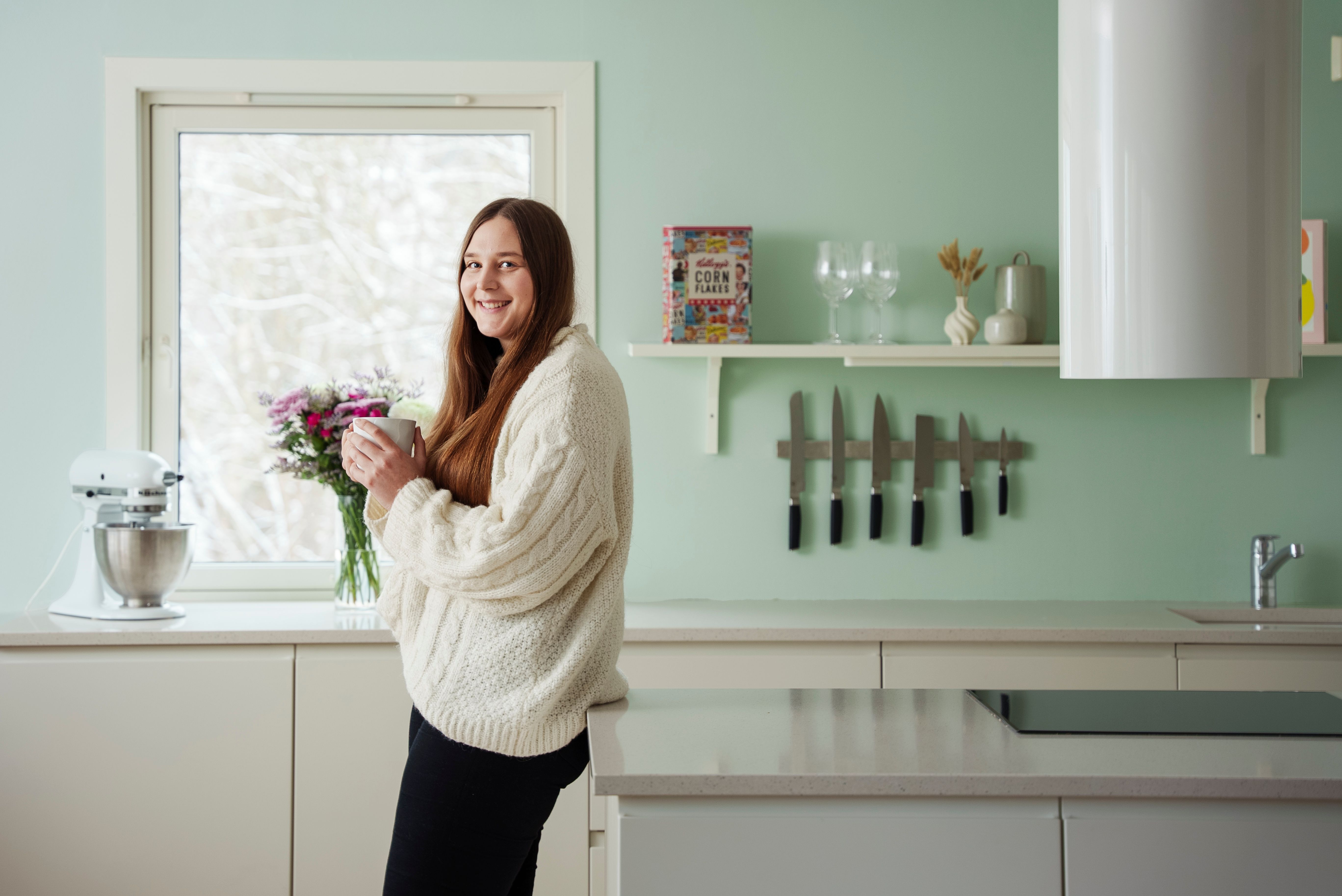 Kristine Istad står i et lyst, moderne kjøkken med en kopp i hånden og smiler mot kamera