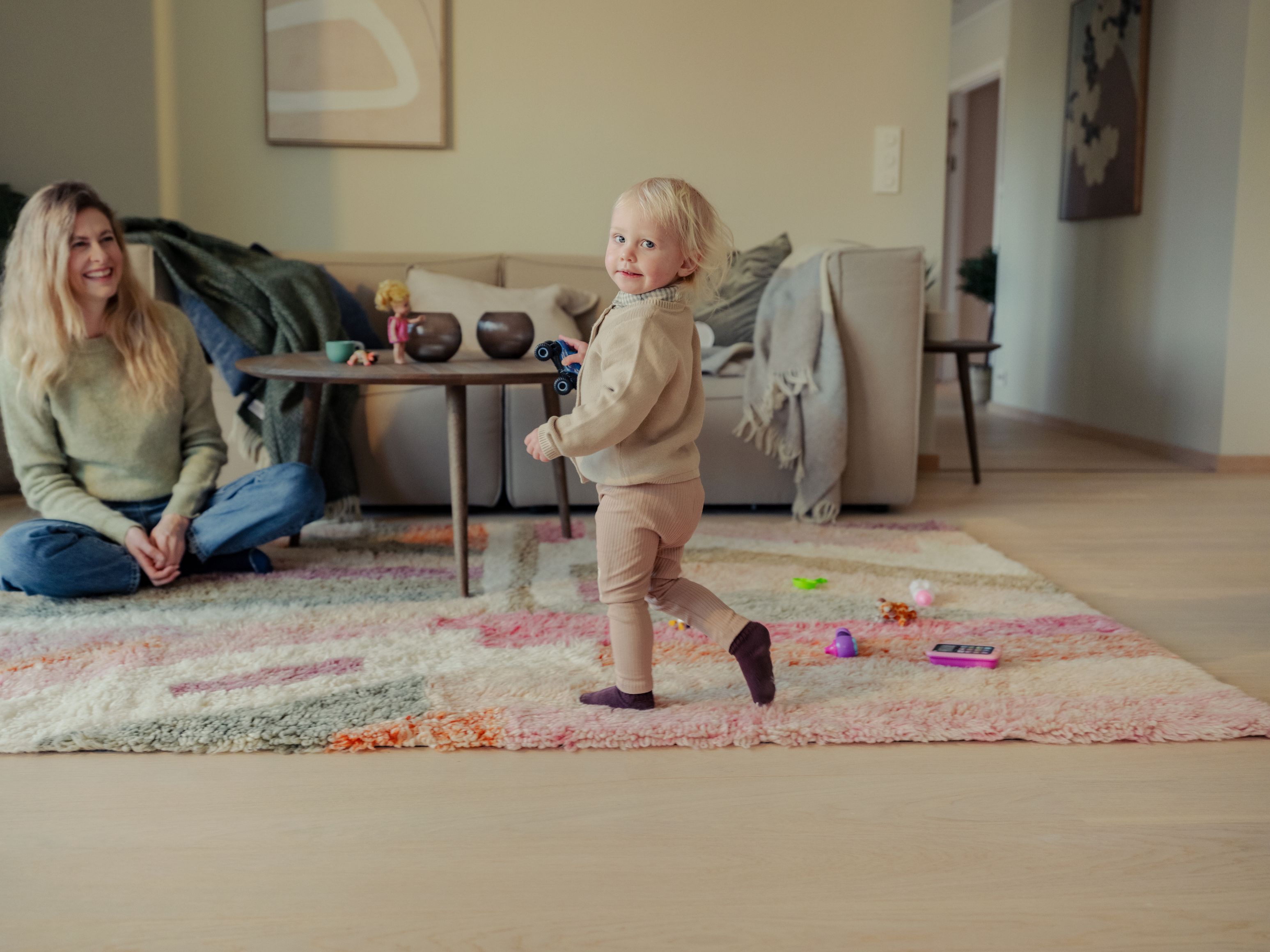 Jente som løper over et teppe og snur seg. Moren sitter på gulvet og ser på henne og smiler.