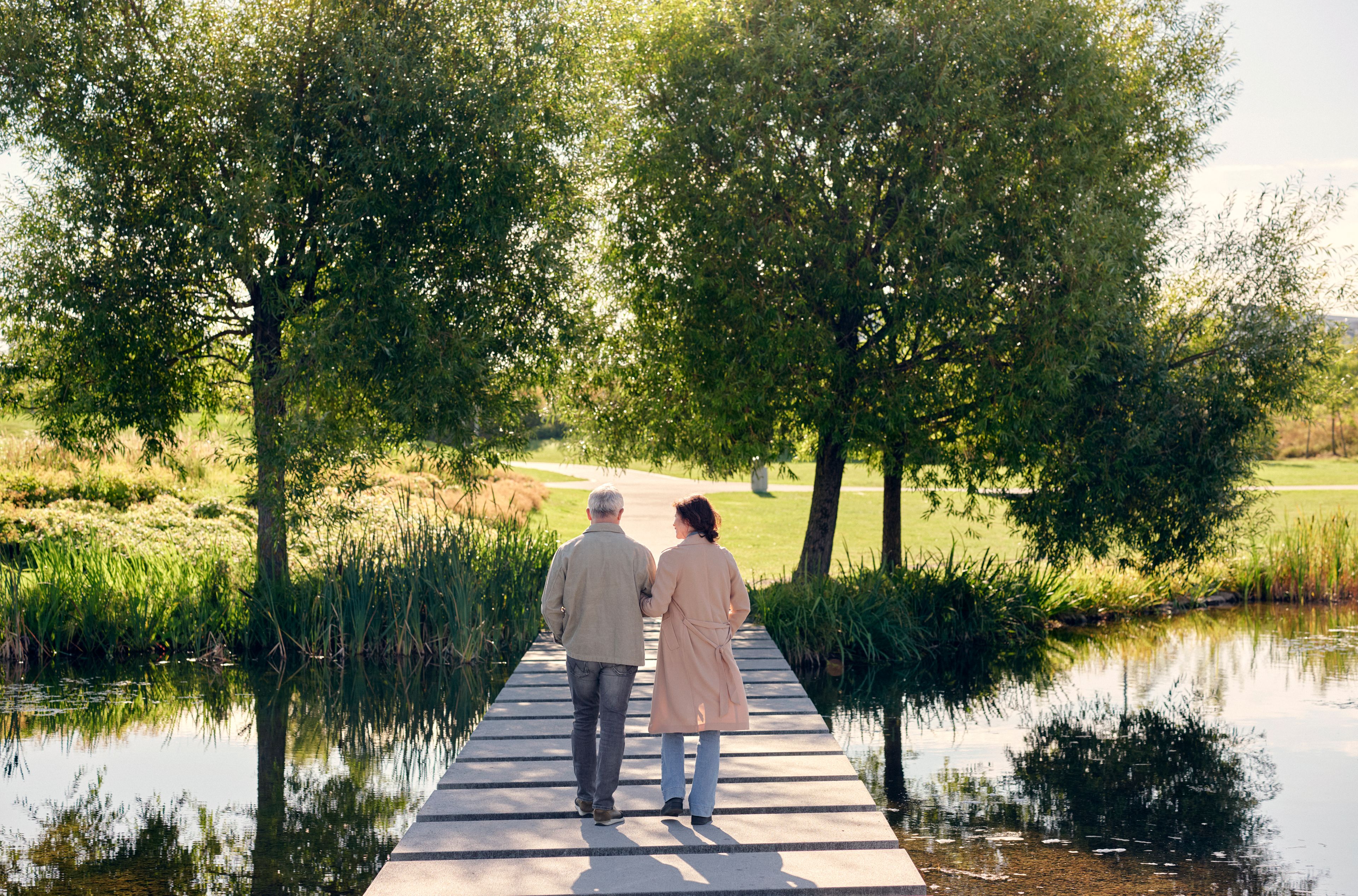 Et par går arm i arm på en fredelig sti over et reflekterende tjern, omgitt av frodige grønne trær og gress, som fremkaller ro og samhørighet.