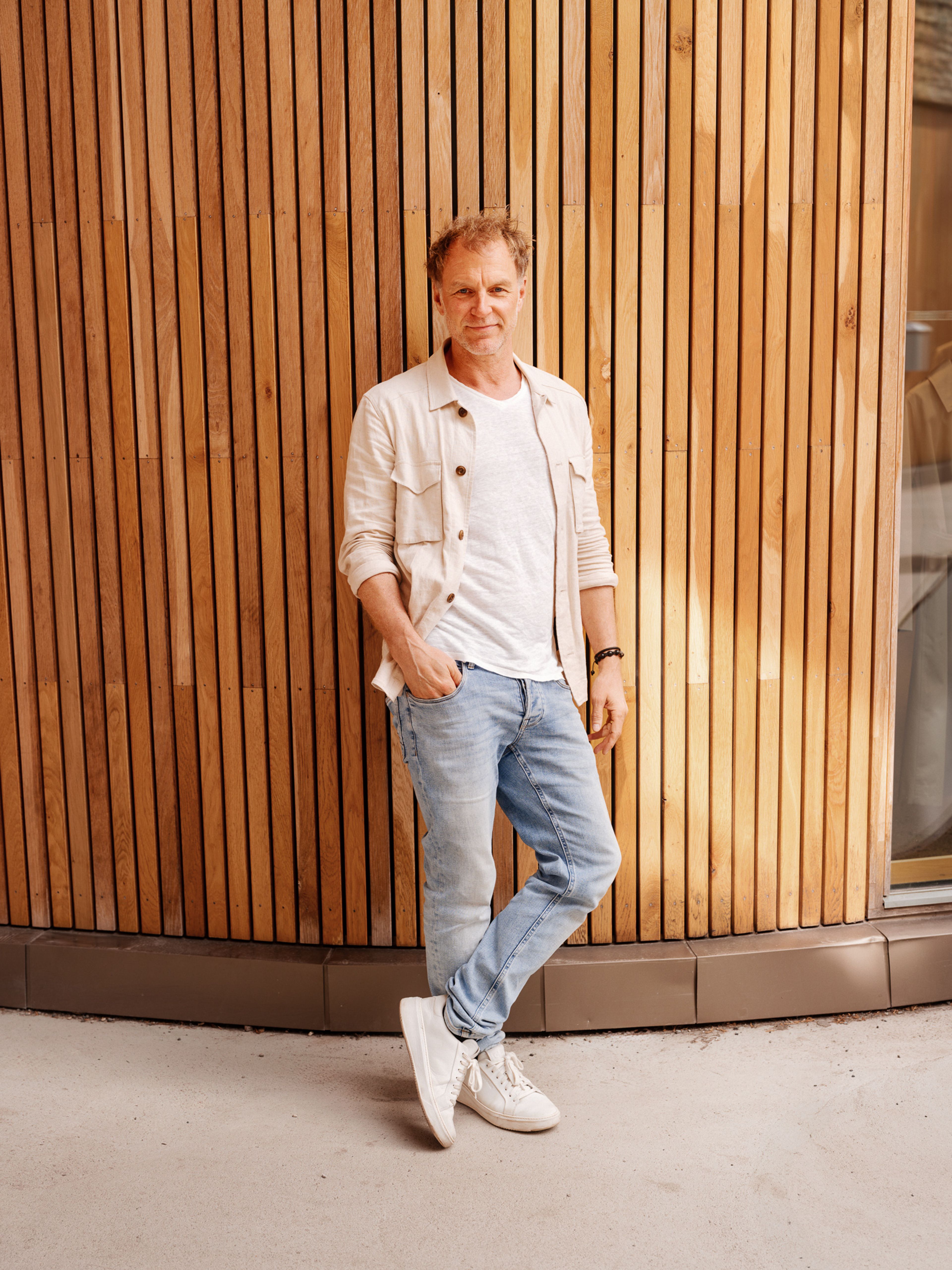 Hans Petter Aaserud har på seg en lys jeans, hvit t-skjorte og kremhvit overshirt. Han lener seg inntil en spilevegg.