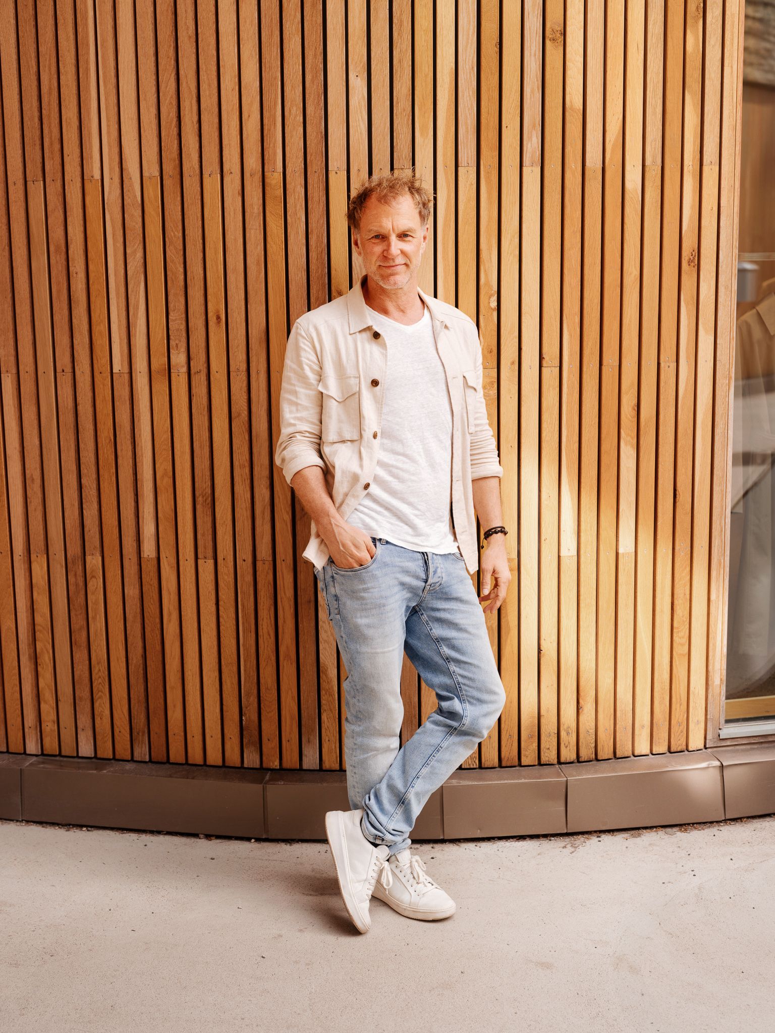 Hans Petter Aaserud har på seg en lys jeans, hvit t-skjorte og kremhvit overshirt. Han lener seg inntil en spilevegg.