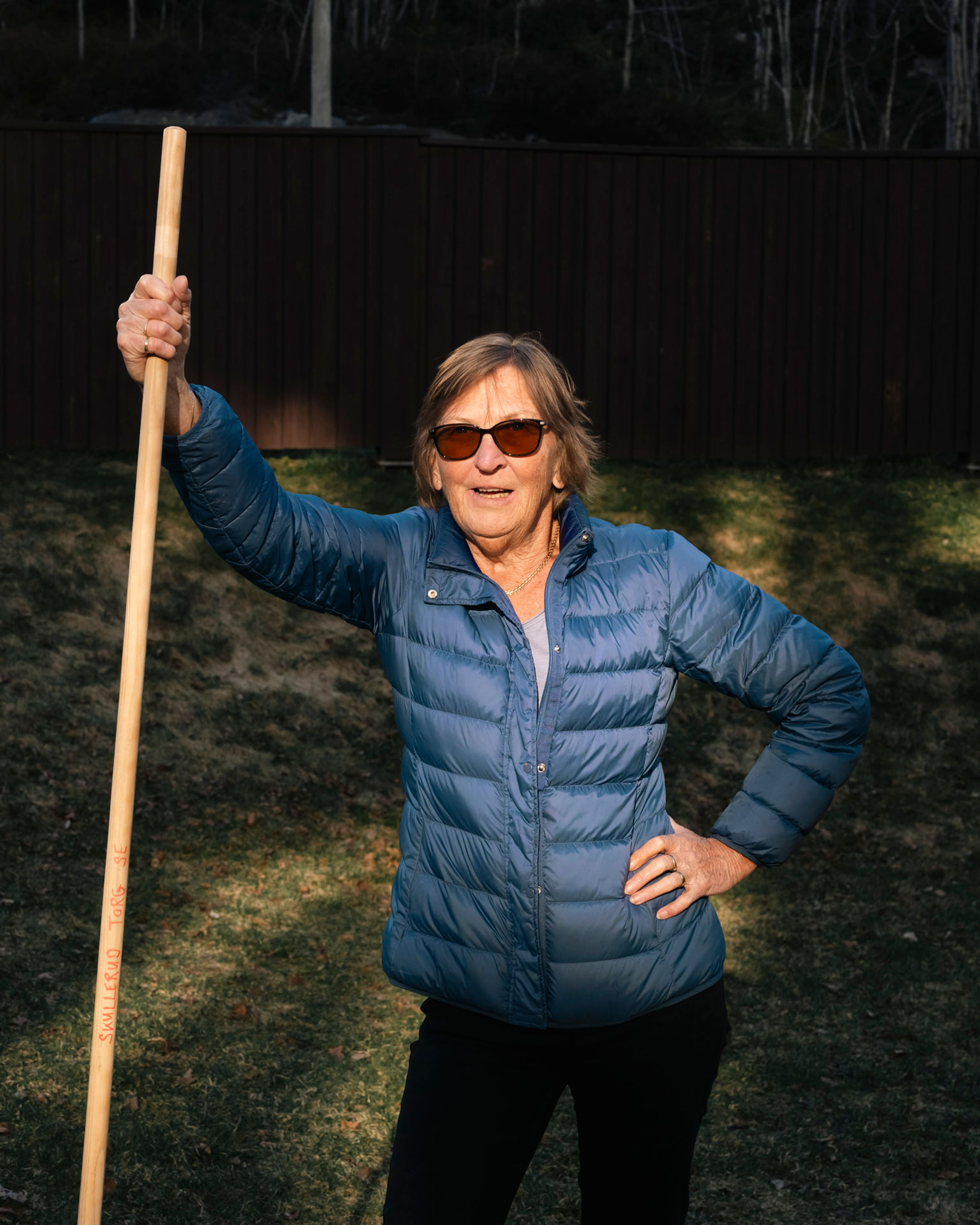 Eldre kvinne i blå dunjakke og solbriller står på plenen og holder et langt riveskaft merket Skullerud Torg.