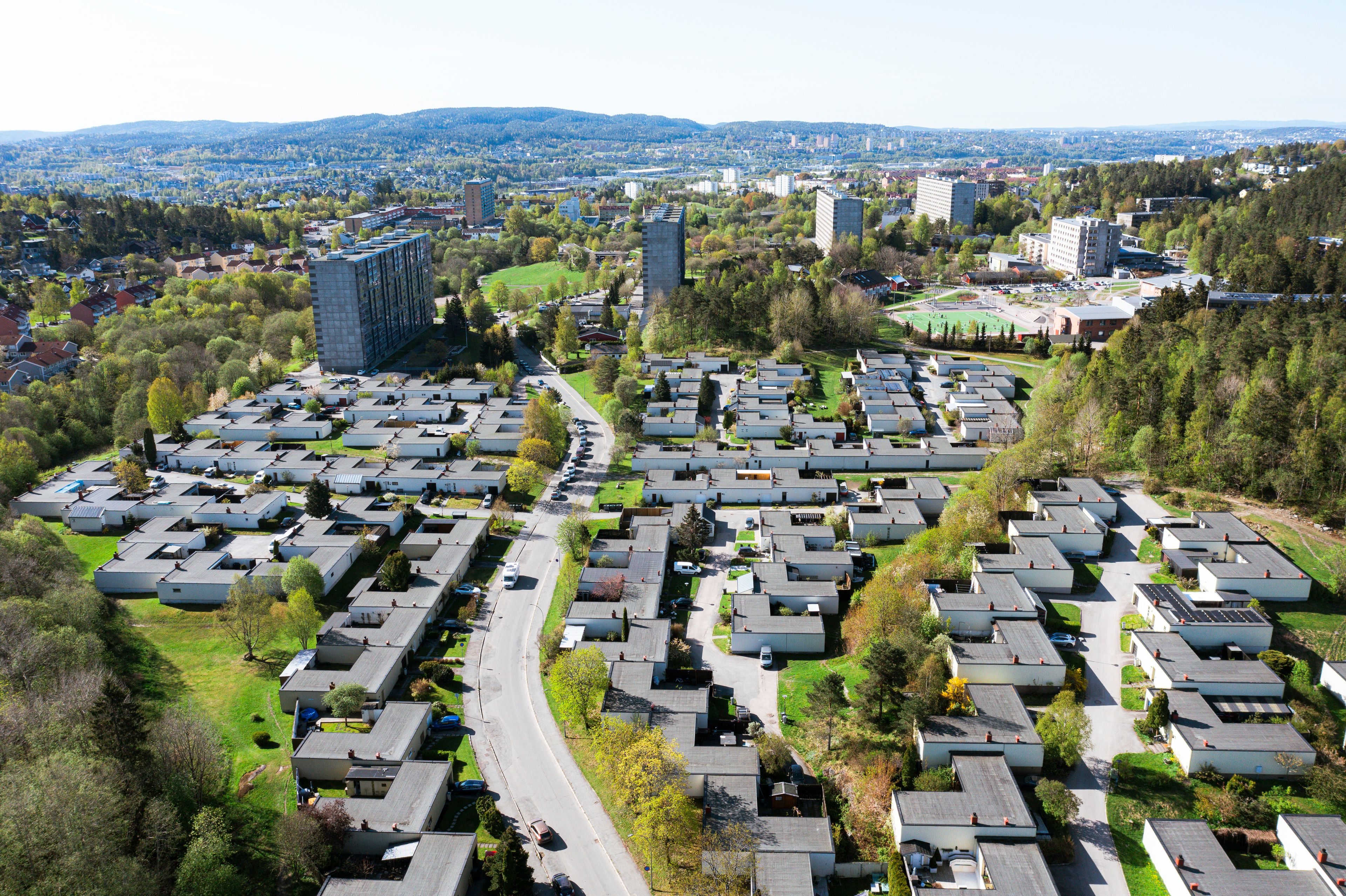 Oversiktsbilde av et stort boligområde med rekkehus og høyblokker, omgitt av trær og grønt landskap