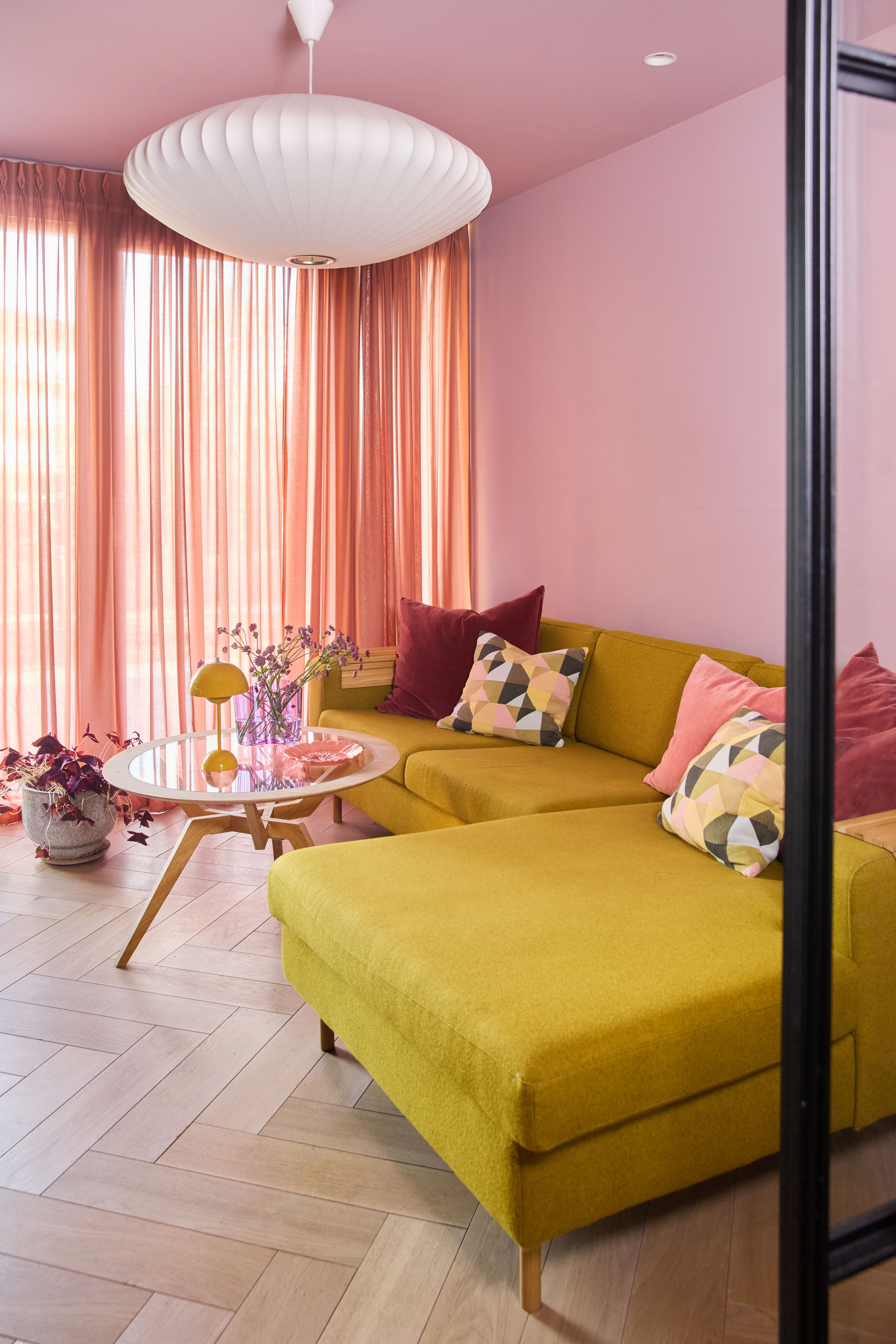 Stue med rosa vegger og tak, sennepsgul hjørnesofa, korallfargede gardiner og hvit Nelson-taklampe i tak.