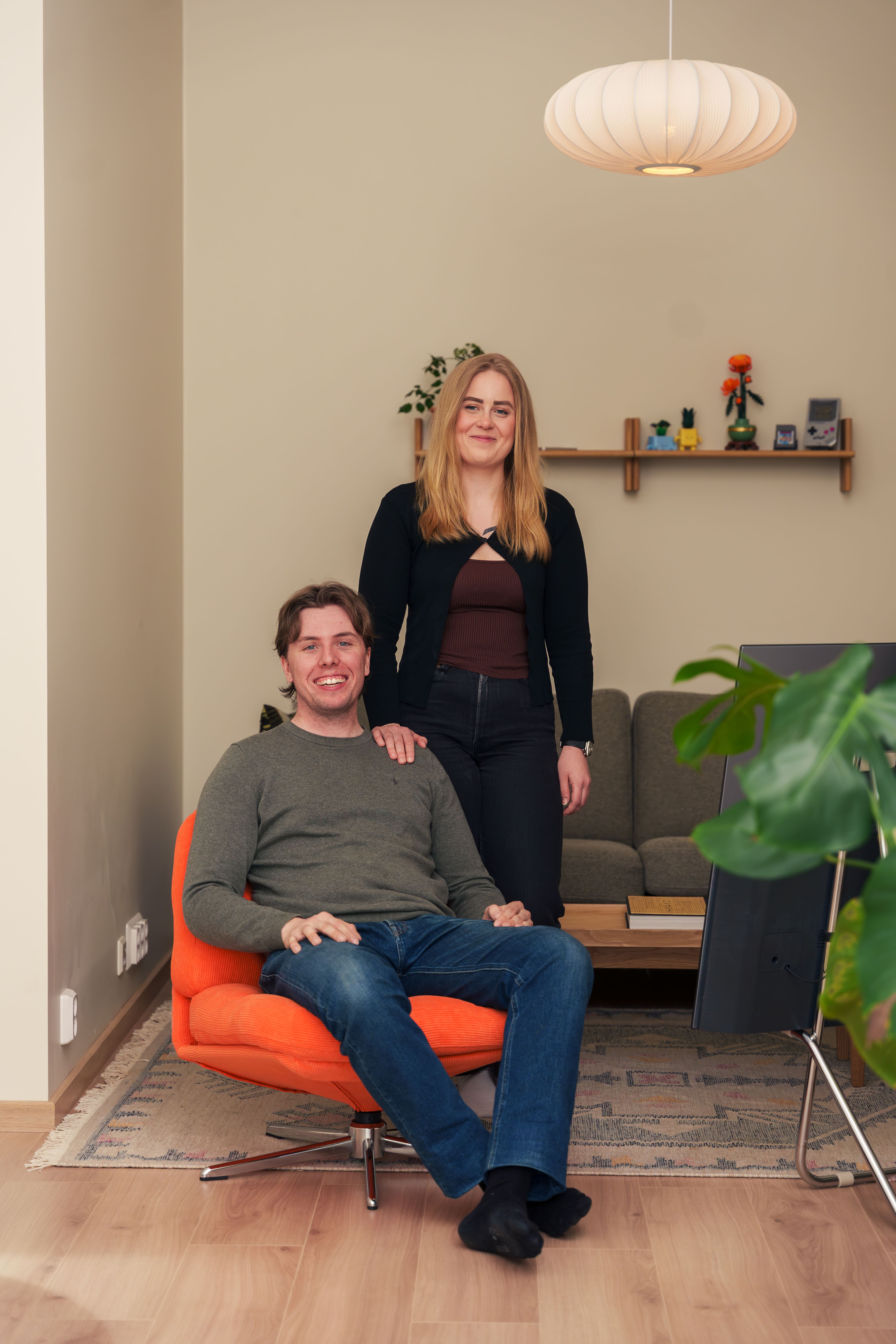 Mann i grønn genser sitter i en oransje dreiestol mens en kvinne står bak ham, under en rund papirlampe i stua.