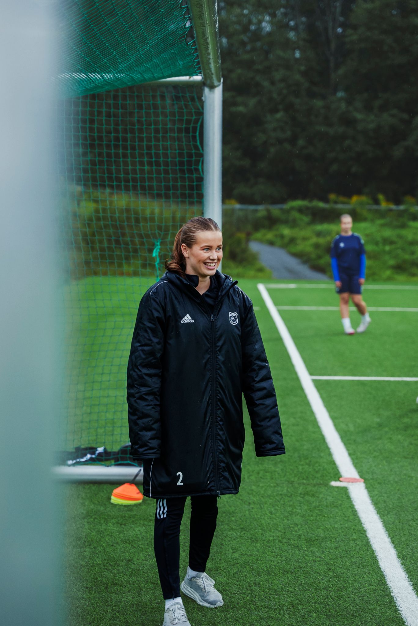 Assistenttrener Hedda smiler og ser mot spillerne som trener ute av bildet på fotballbanen.