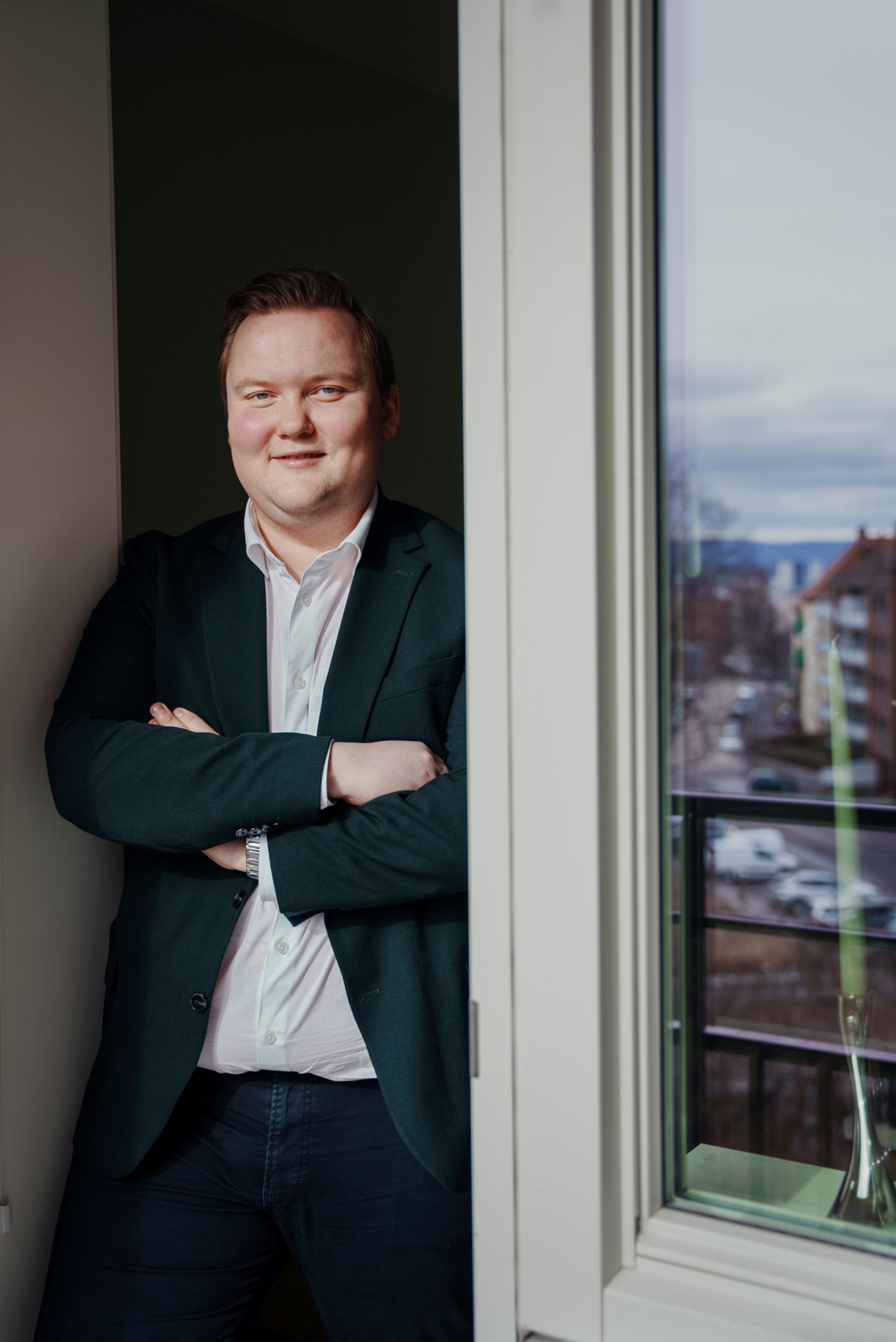 Fredrik Campbell Pedersen står kledd i dress med armene i kors i en døråpning.