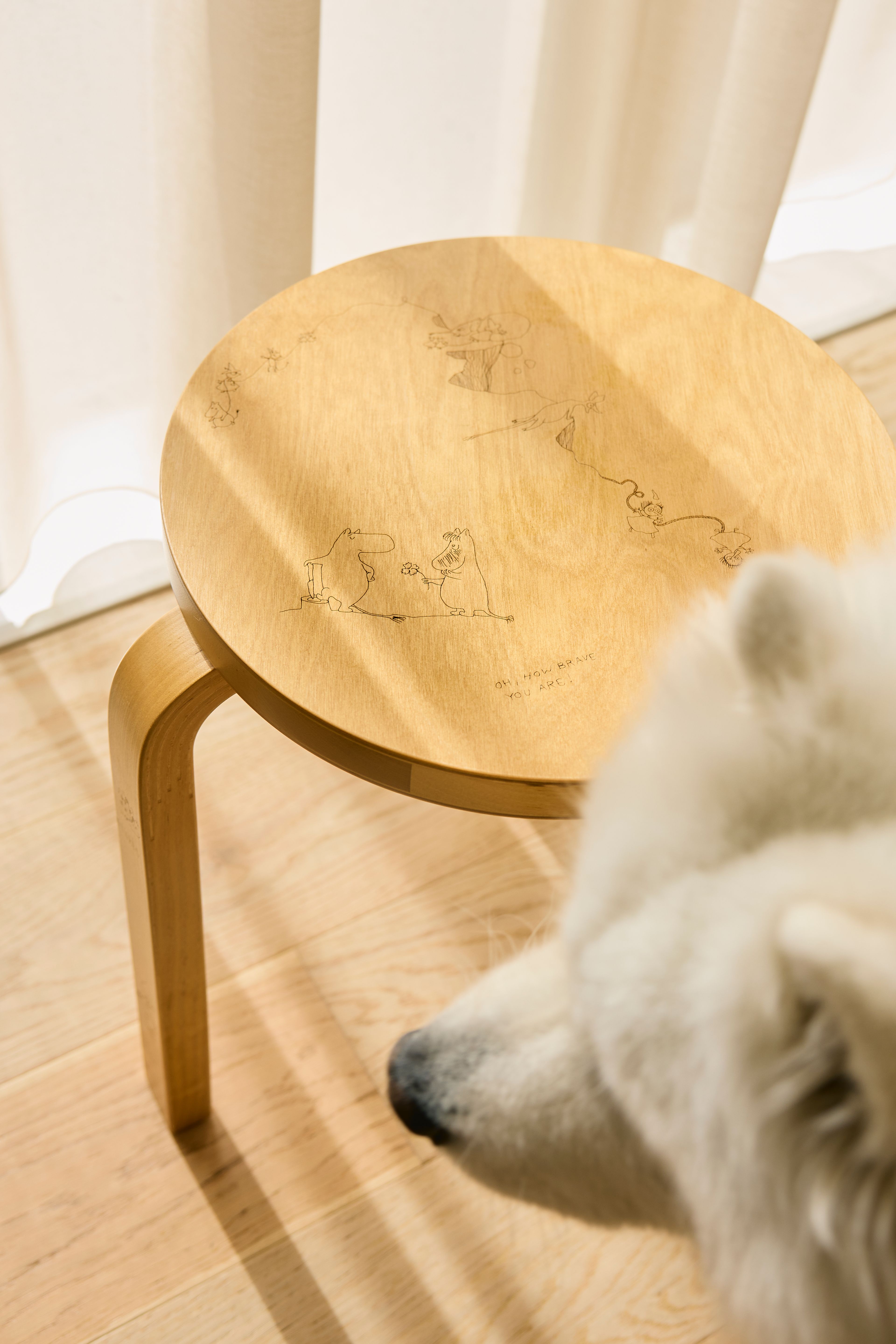 Rund krakk i lyst bjørkefiner med inngraverte Mummi-figurer på setet, med snuten til en hvit hund i forgrunnen.