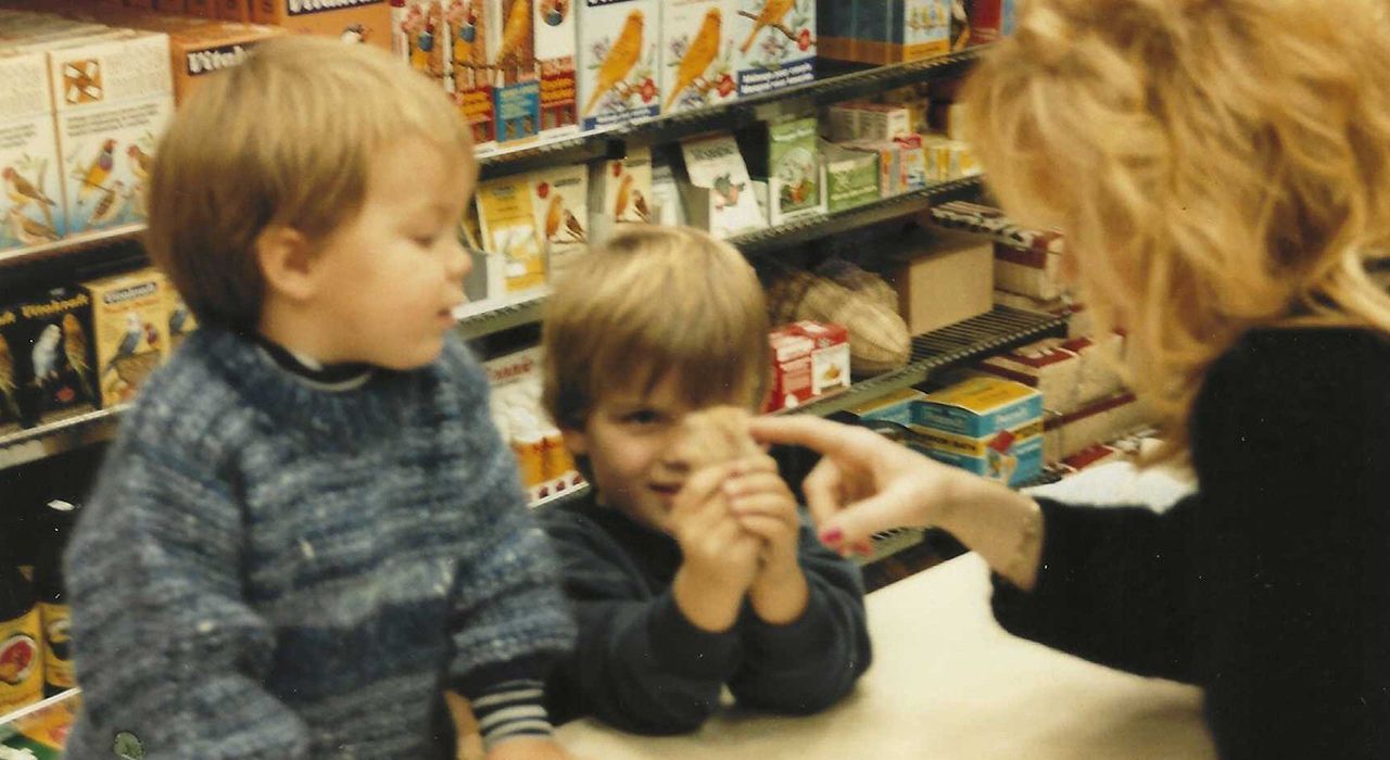 Foto av Frode og Morten Juul som barn. I bildet står de bak disken i dyrebutikken Dyrego og snakker med en dame.