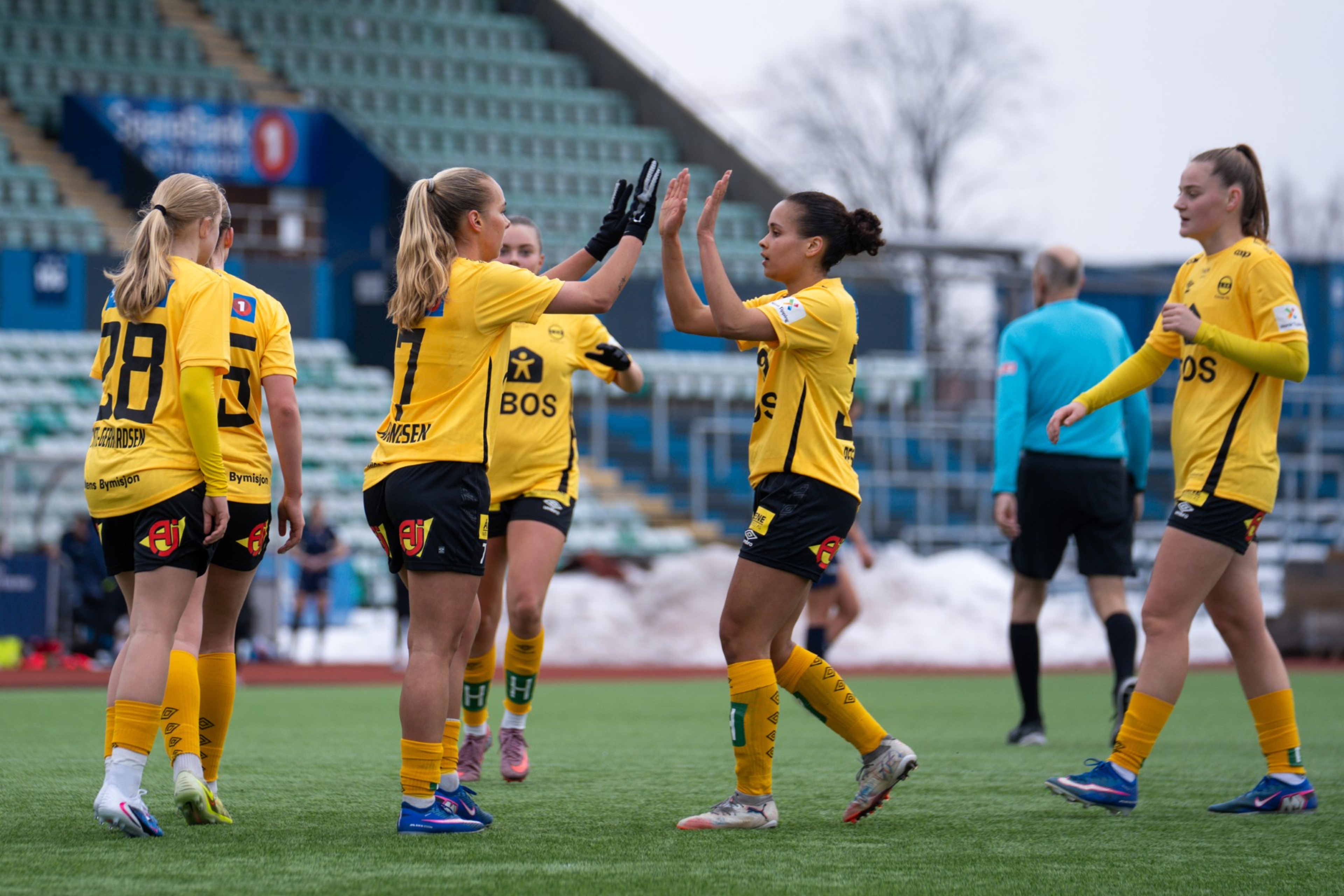 Foto av seks kvinnelige fotballspillere for Lillestrøm fotballklubb som er samlet på banen. To av dem tar high five.