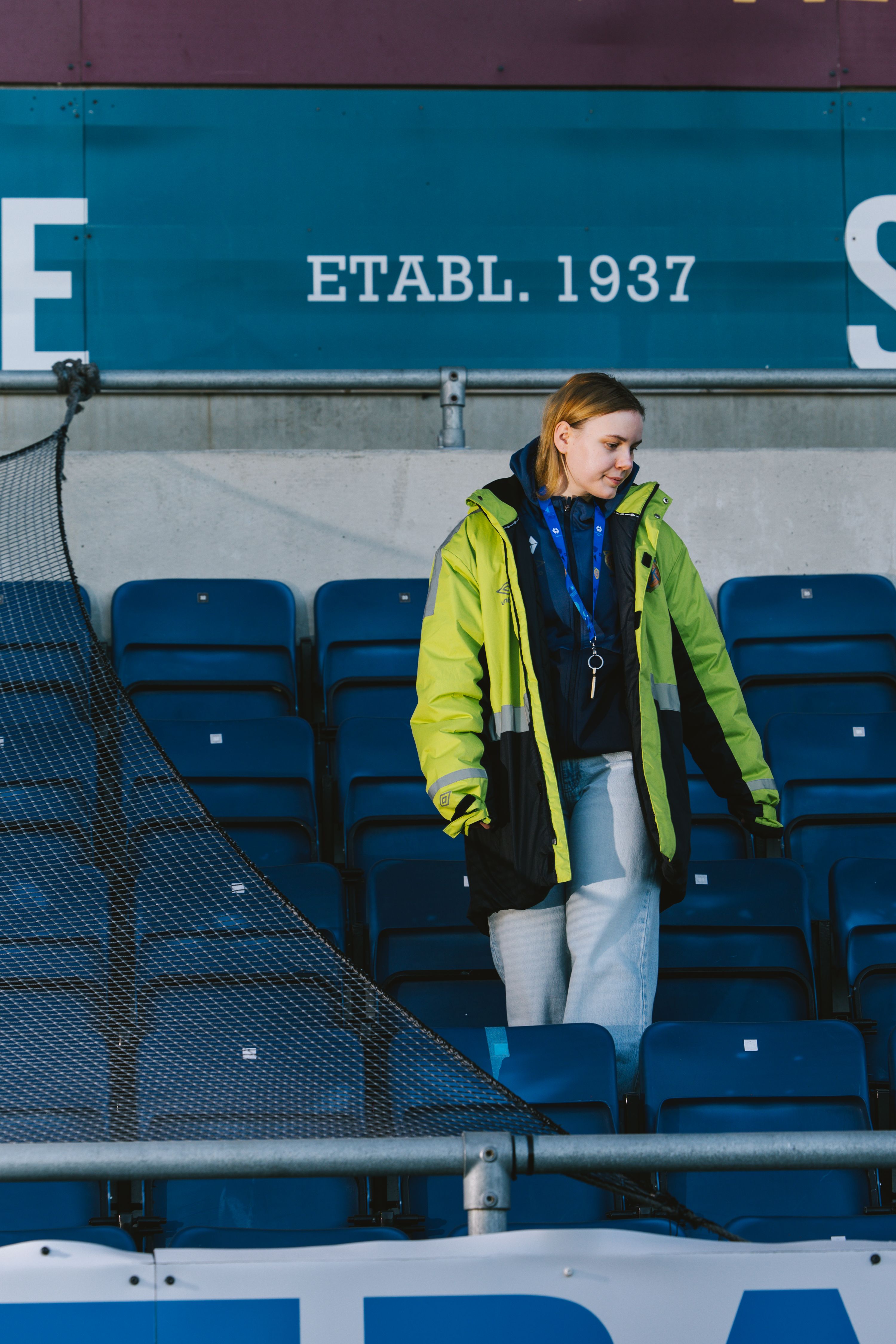 Kristina står mellom de tomme blå setene i tribunen, under det historiske skiltet «ETABL. 1937» på Aalesunds stadion.