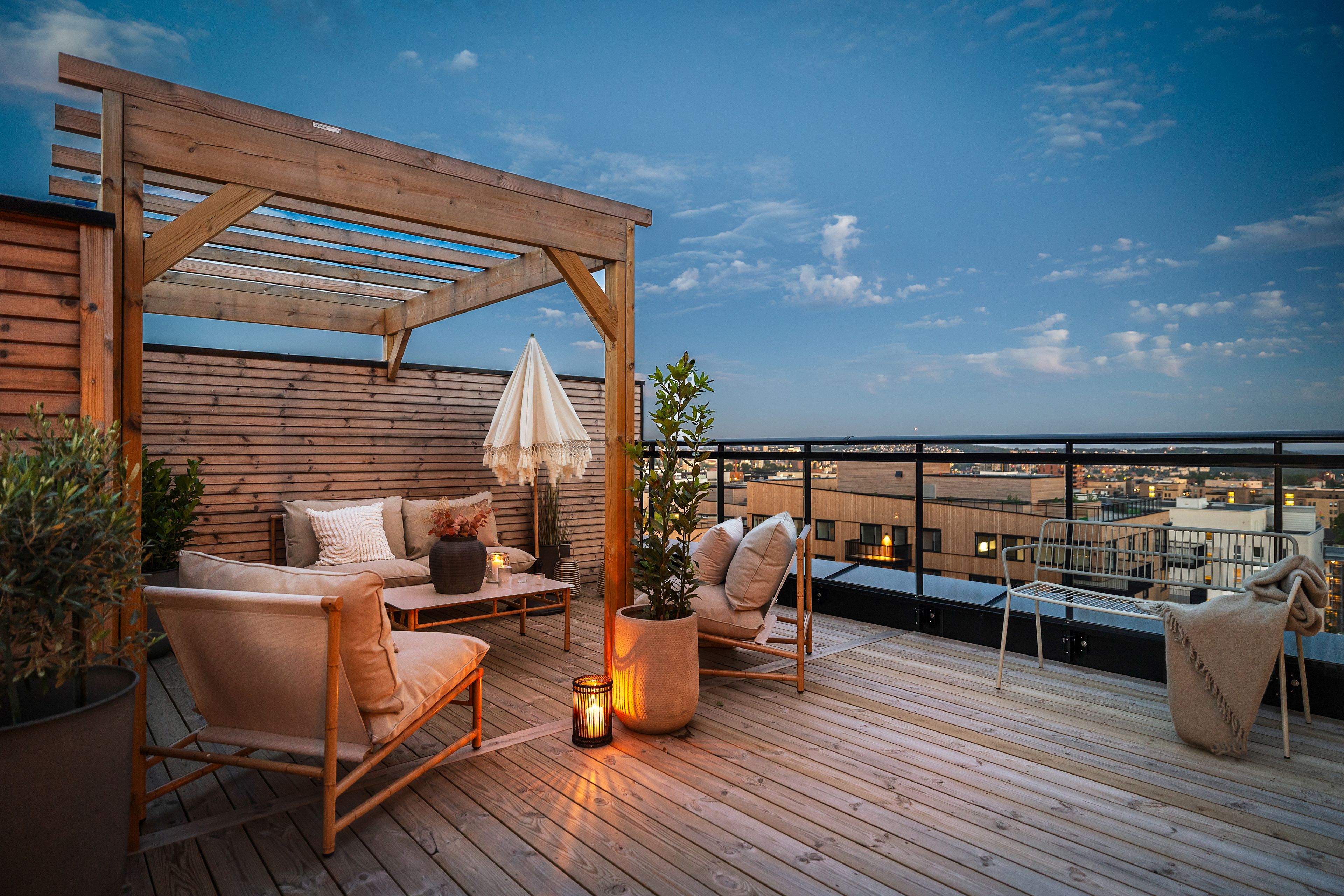 Kveldsbilde av takterrasse i Løren botaniske med sofagruppe under pergola i tre. Utsikt mot by og åser i bakgrunnen.
