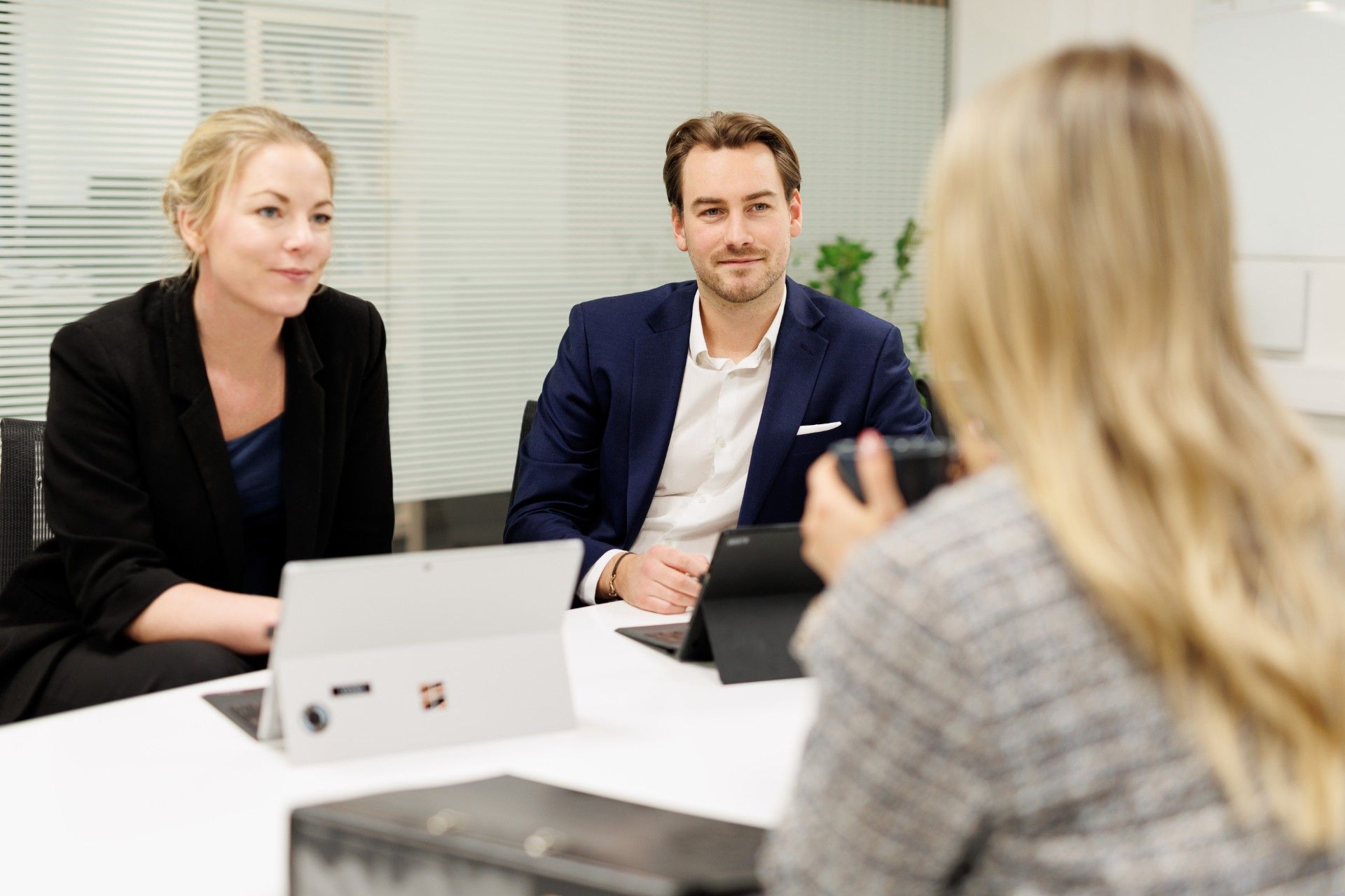 Tre personer sitter ved et bord. To personer sitter ved siden av hverandre og hjelper en over bordet. Han har på seg dress.
