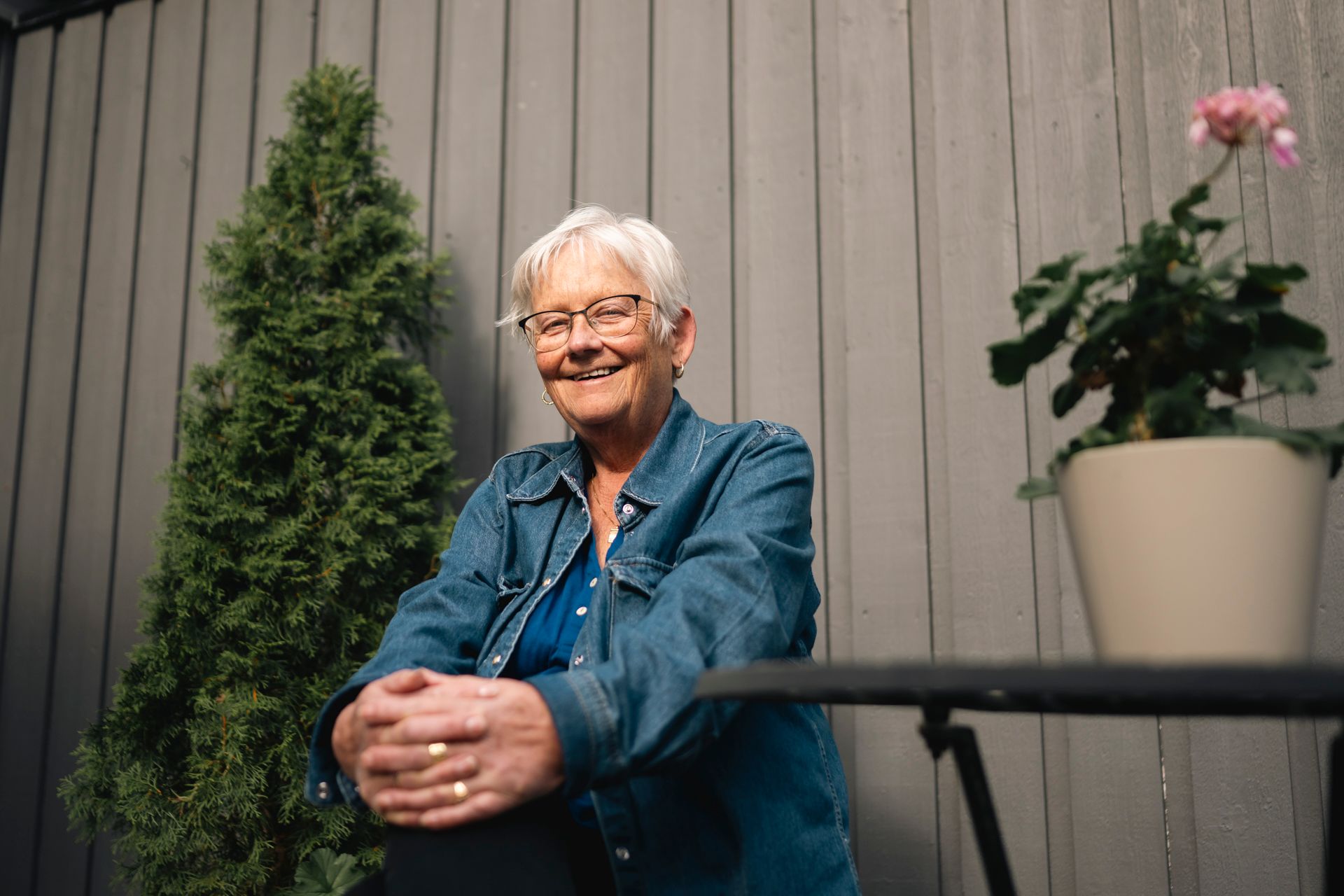 Anne-Berit Snekkersveen sitter ute foran grå kledning, smiler mot kamera med hendene hvilende på knærne.