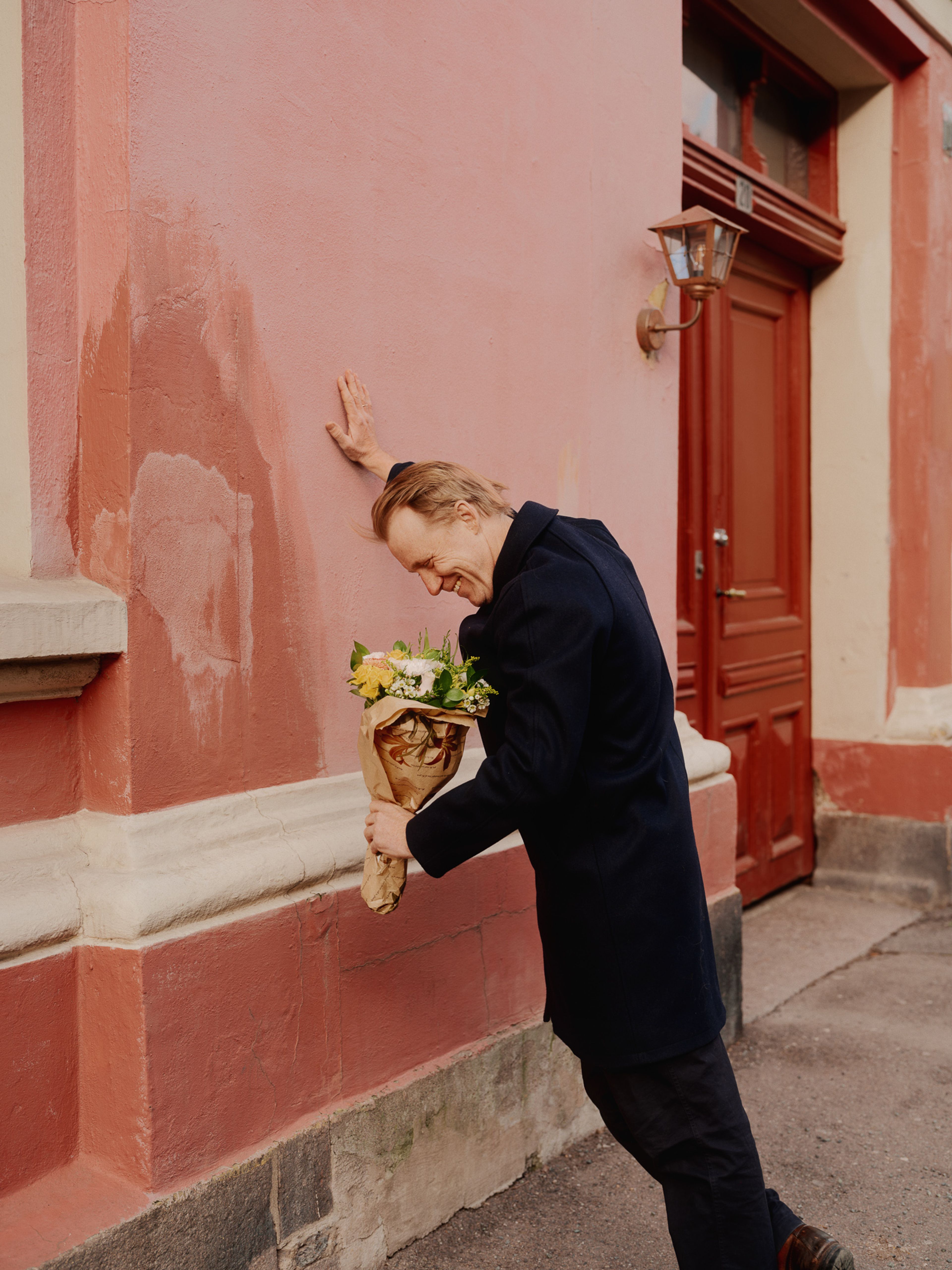 Thorbjørn Harr holder en blomsterbukett i hånda, han lukter på den mens han lener seg inn mot en rosa vegg.