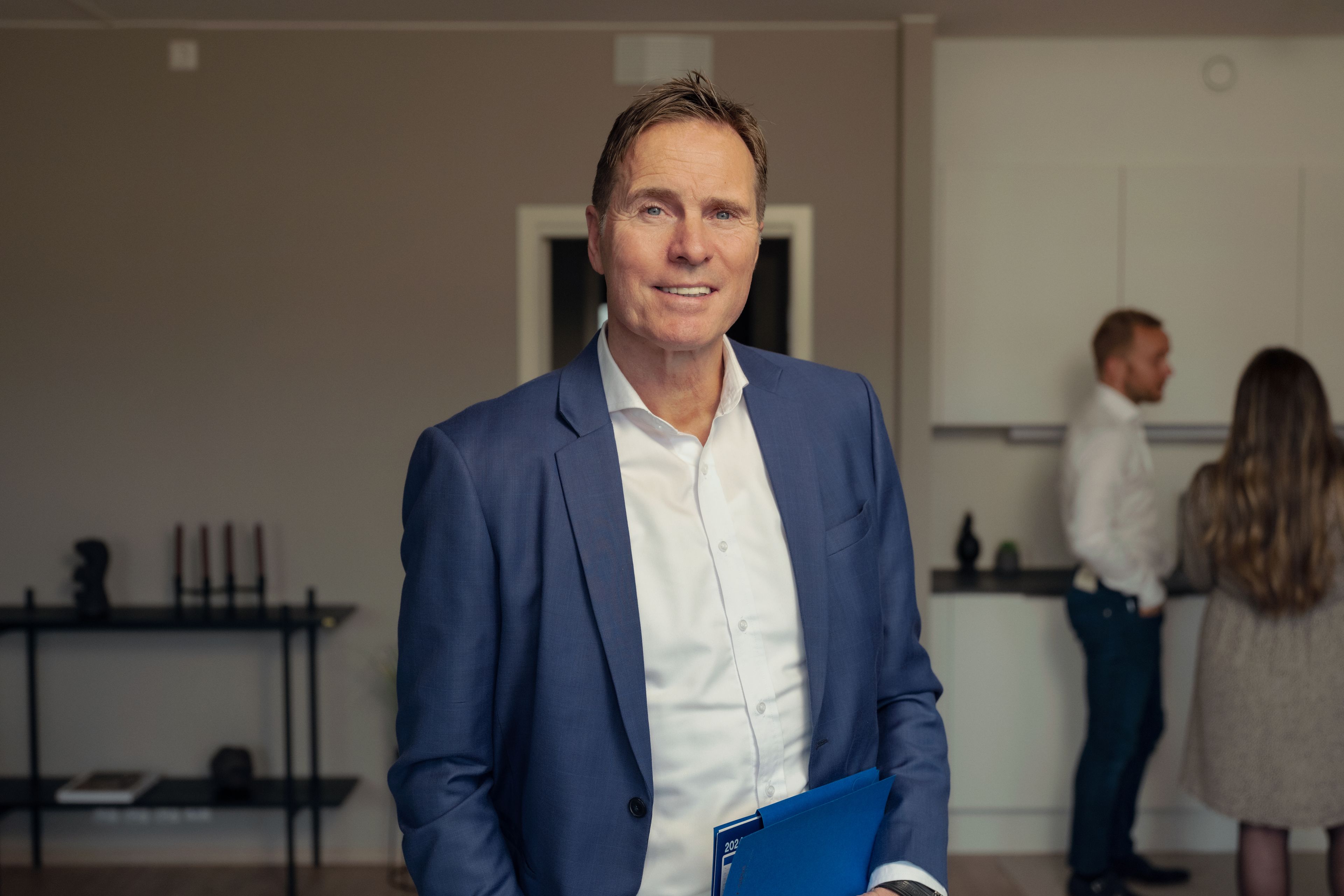 Asbjørn Myrstøl står i en stue og smiler til kamera, med et prospekt i hånden.