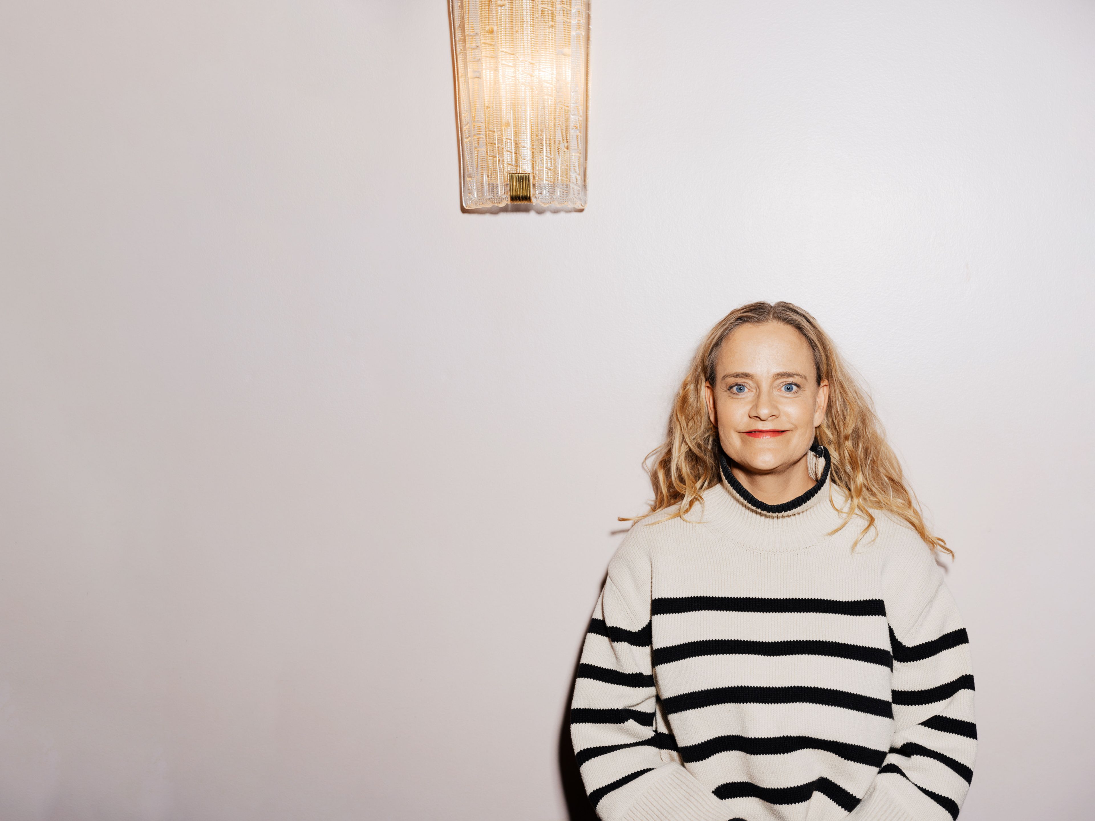 Foto av Henriette Steenstrup inntil en hvit vegg med en lampe hengende på veggen