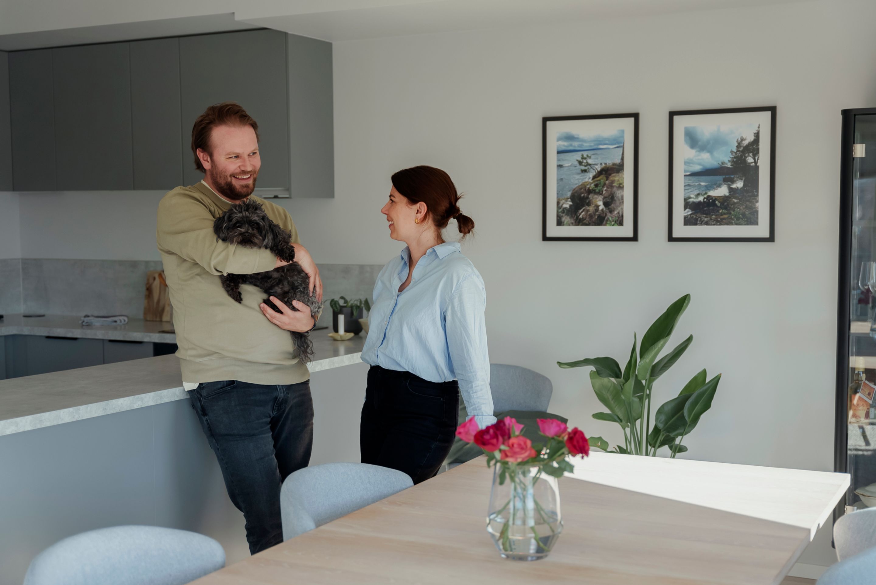 Martin står og bærer hunden Joa ved siden av Stine. De står lent mot kjøkkenbenken i spisestuen, som er kledd i lyse farger.