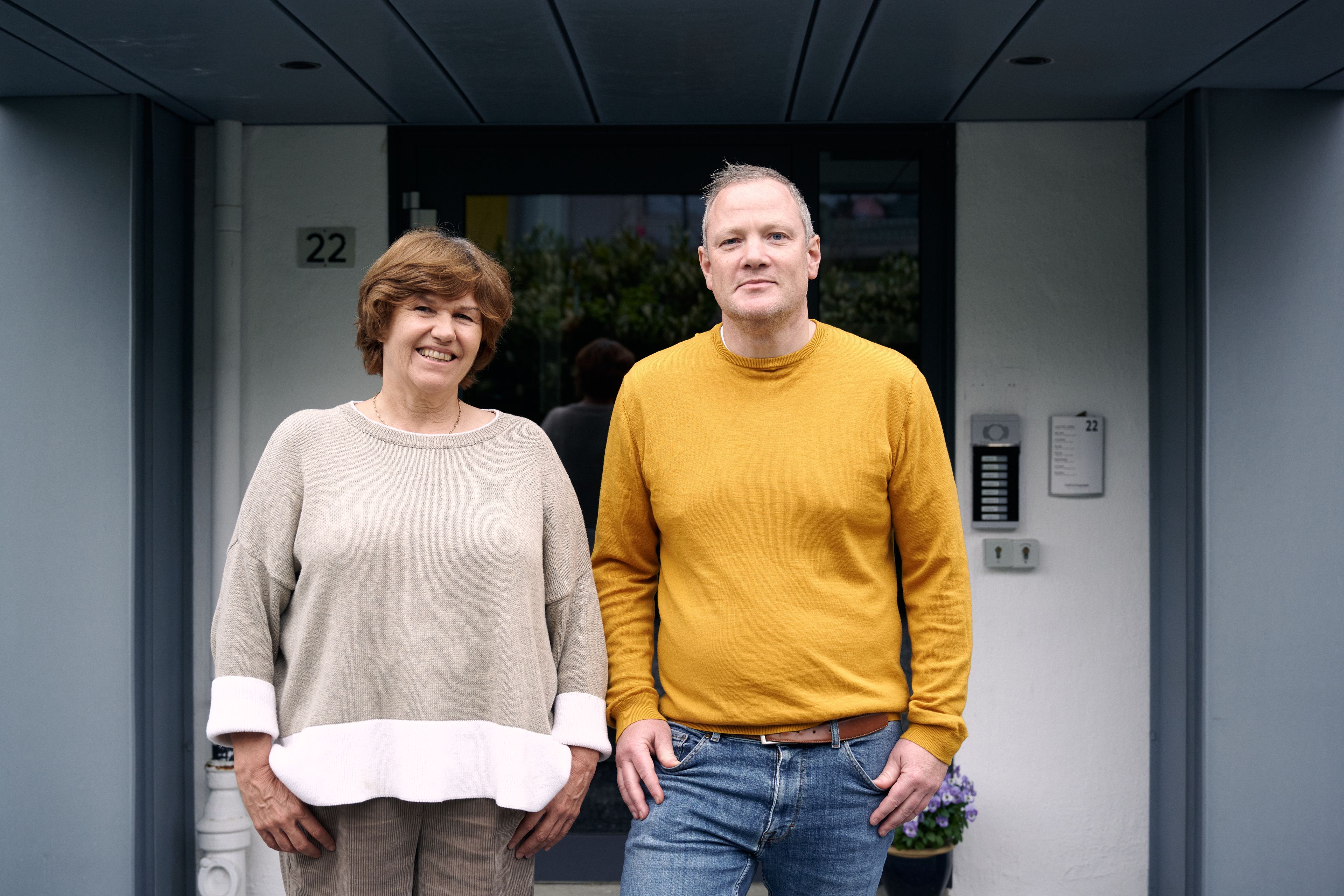 Kvinne i beige genser og mann i gul genser står ved siden av hverandre foran en inngangsdør merket 22.