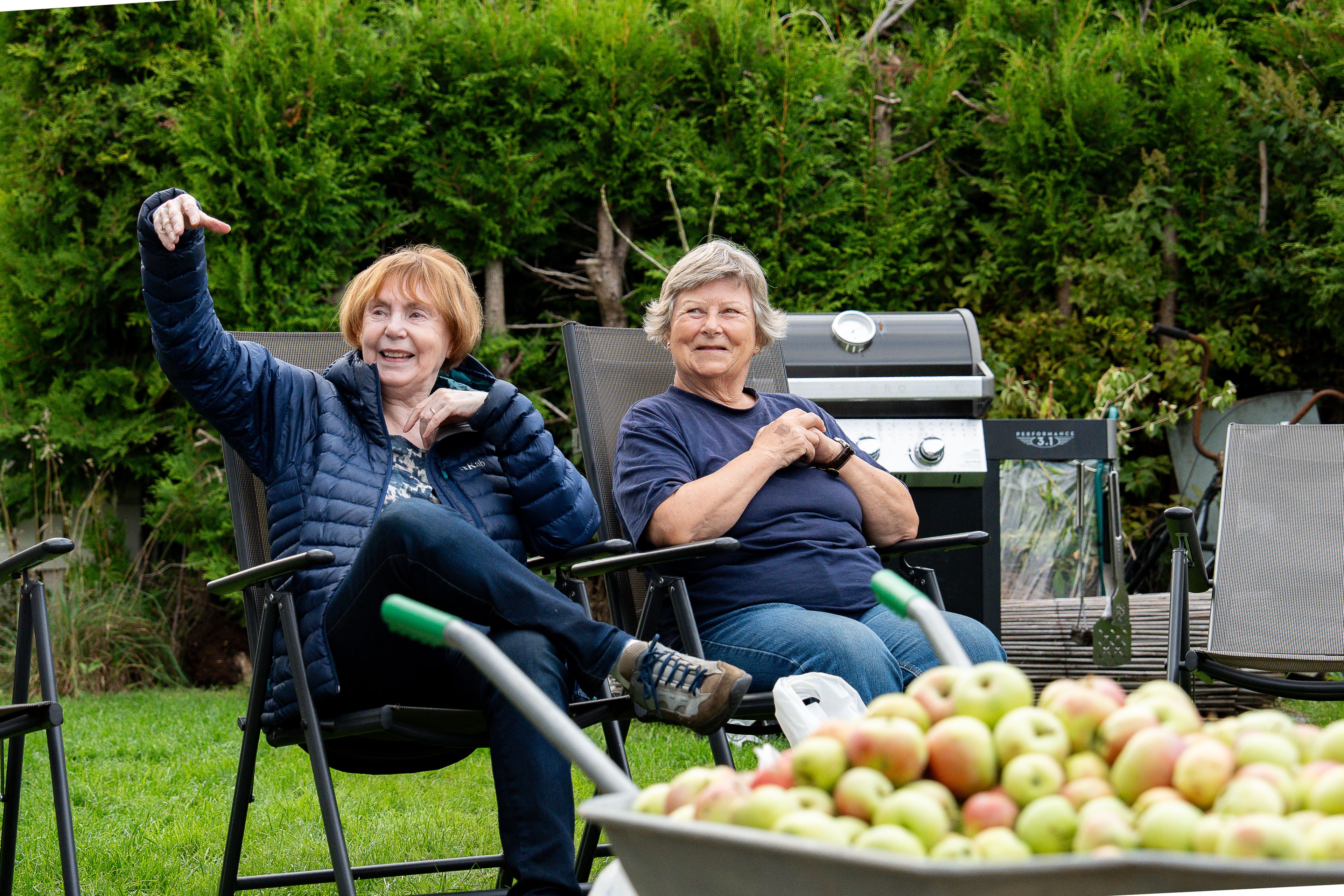 To voksne kvinner sitter i to stoler i en hage og smiler, med epler i forgrunnen