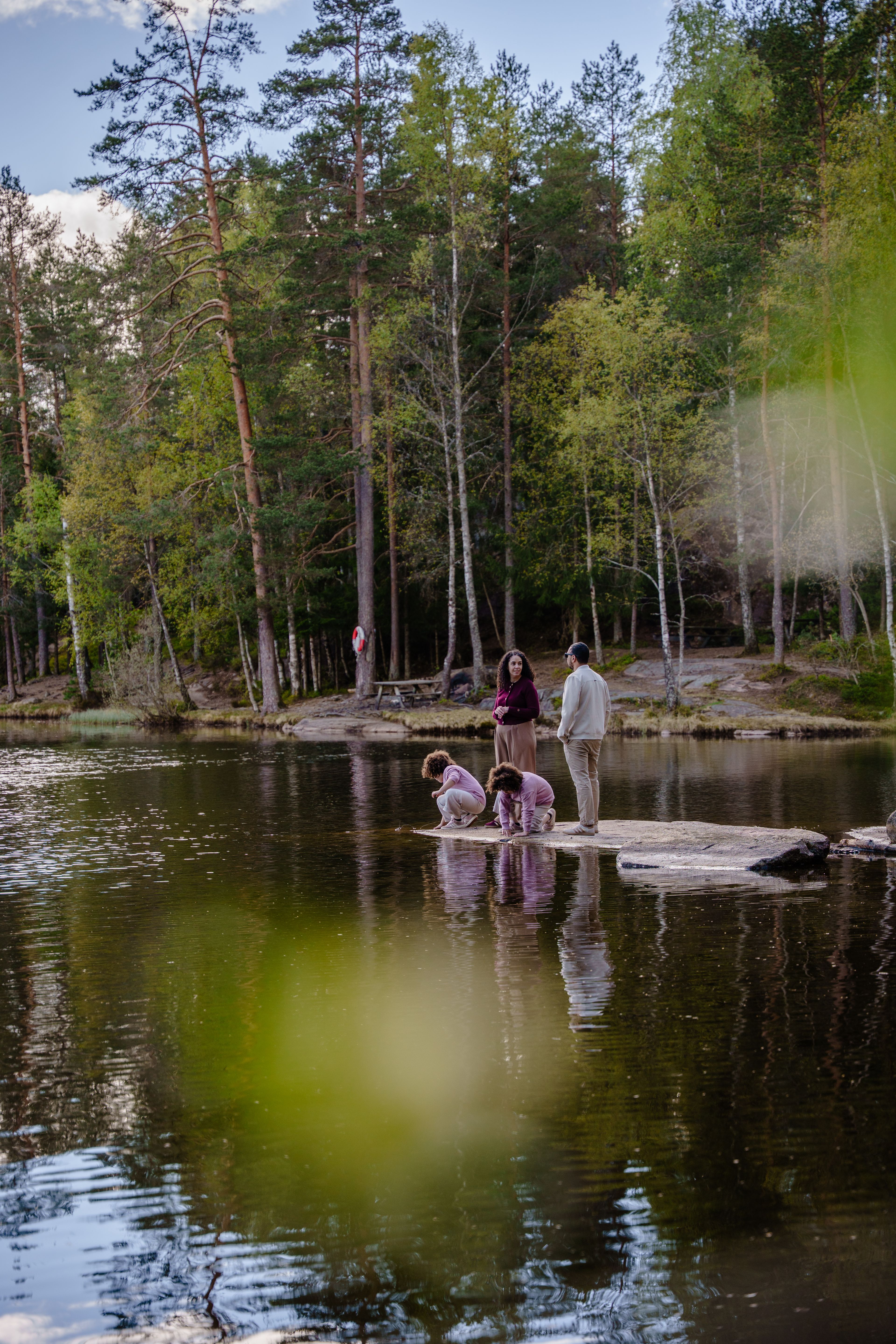 En familie på fire står på et svaberg ved et stille skogsvann. De to barna sitter på huk ved vannkanten.