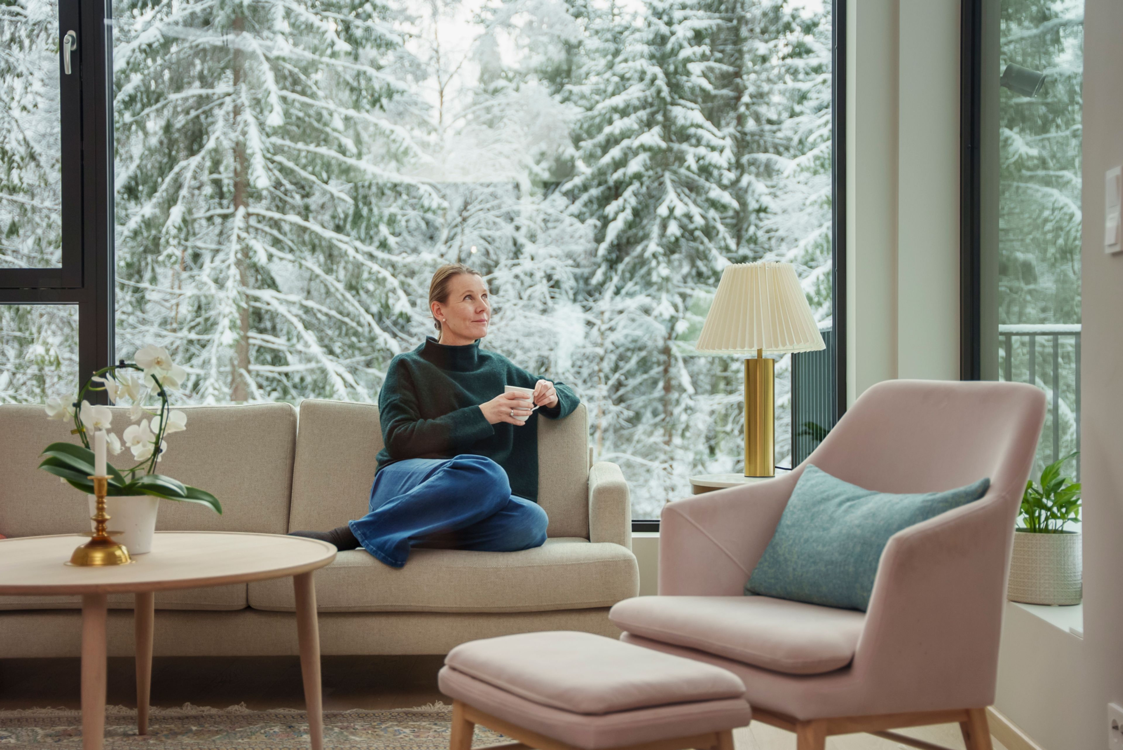 Nina sitter i sofaen sin i huset på Vitigrend. Utenfor ser du at hun er tett på naturen og snøkledde trør.