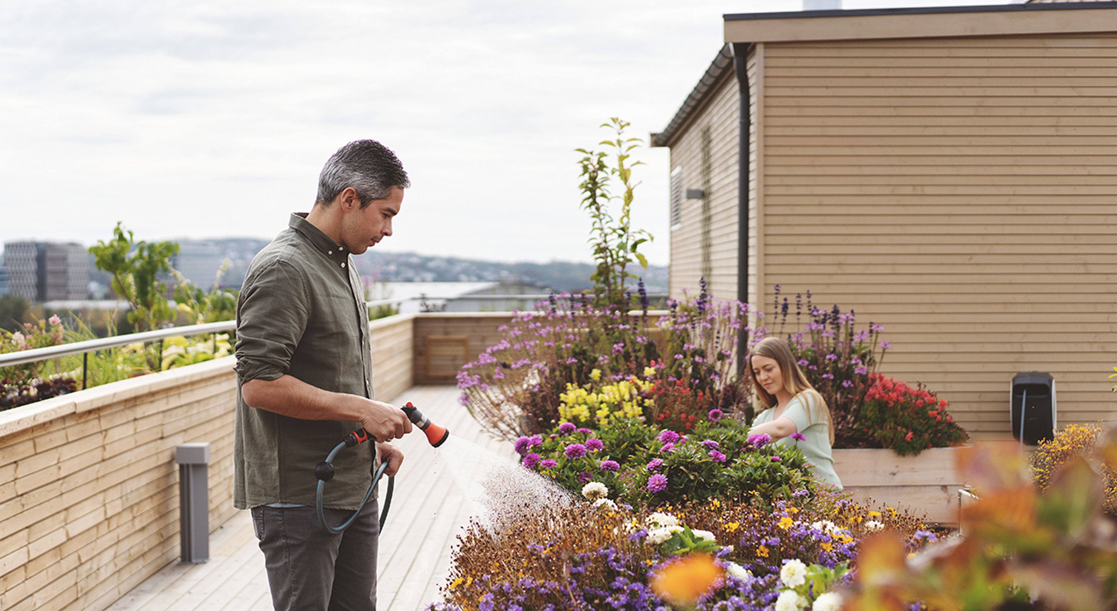 En mann som vanner blomster, mens en dame planter blomster på en takterrasse på Ulven.