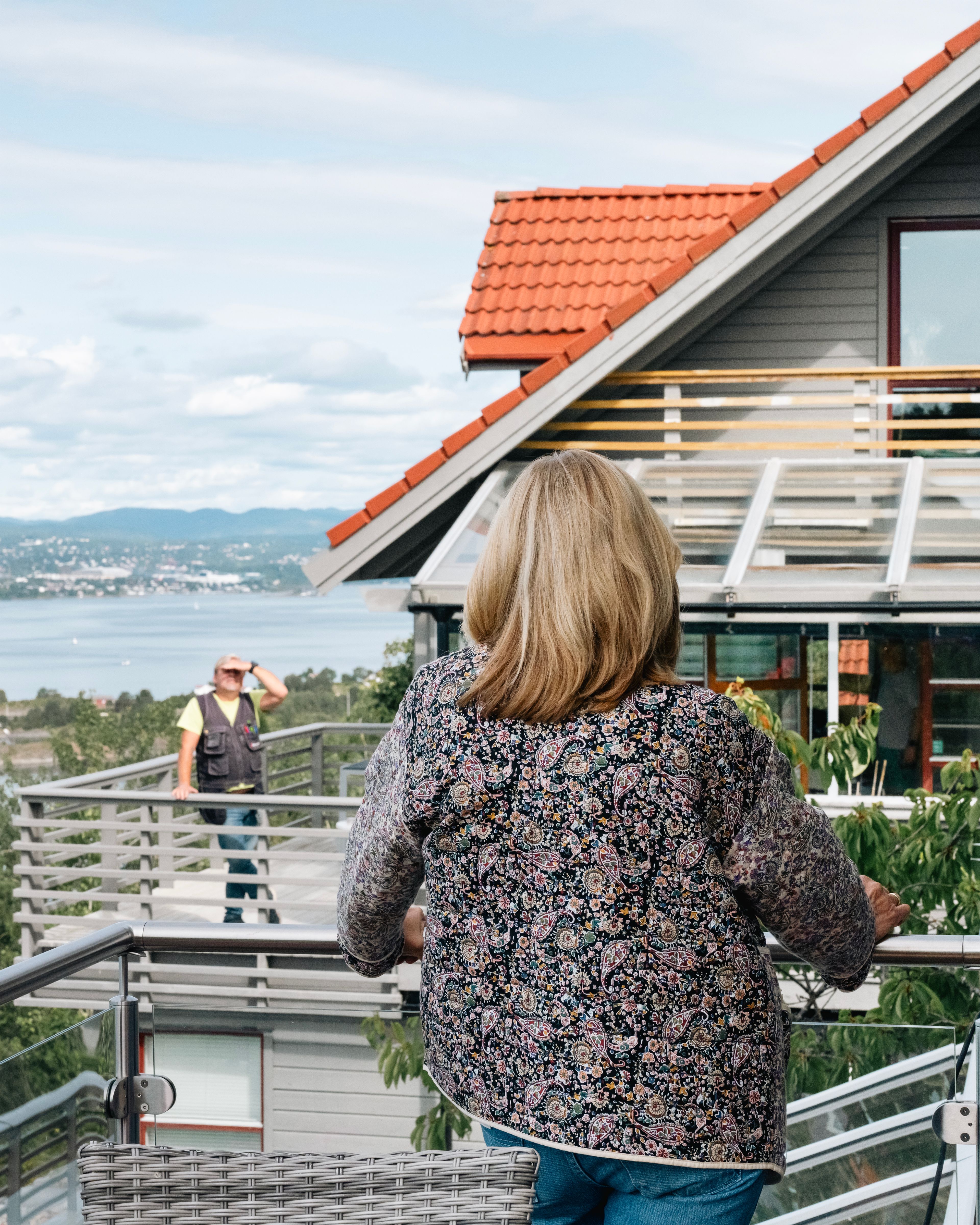 En dame hilser på en mann over rekkverket i borettslaget. Det er hus i bakgrunnen og du ser vannet.