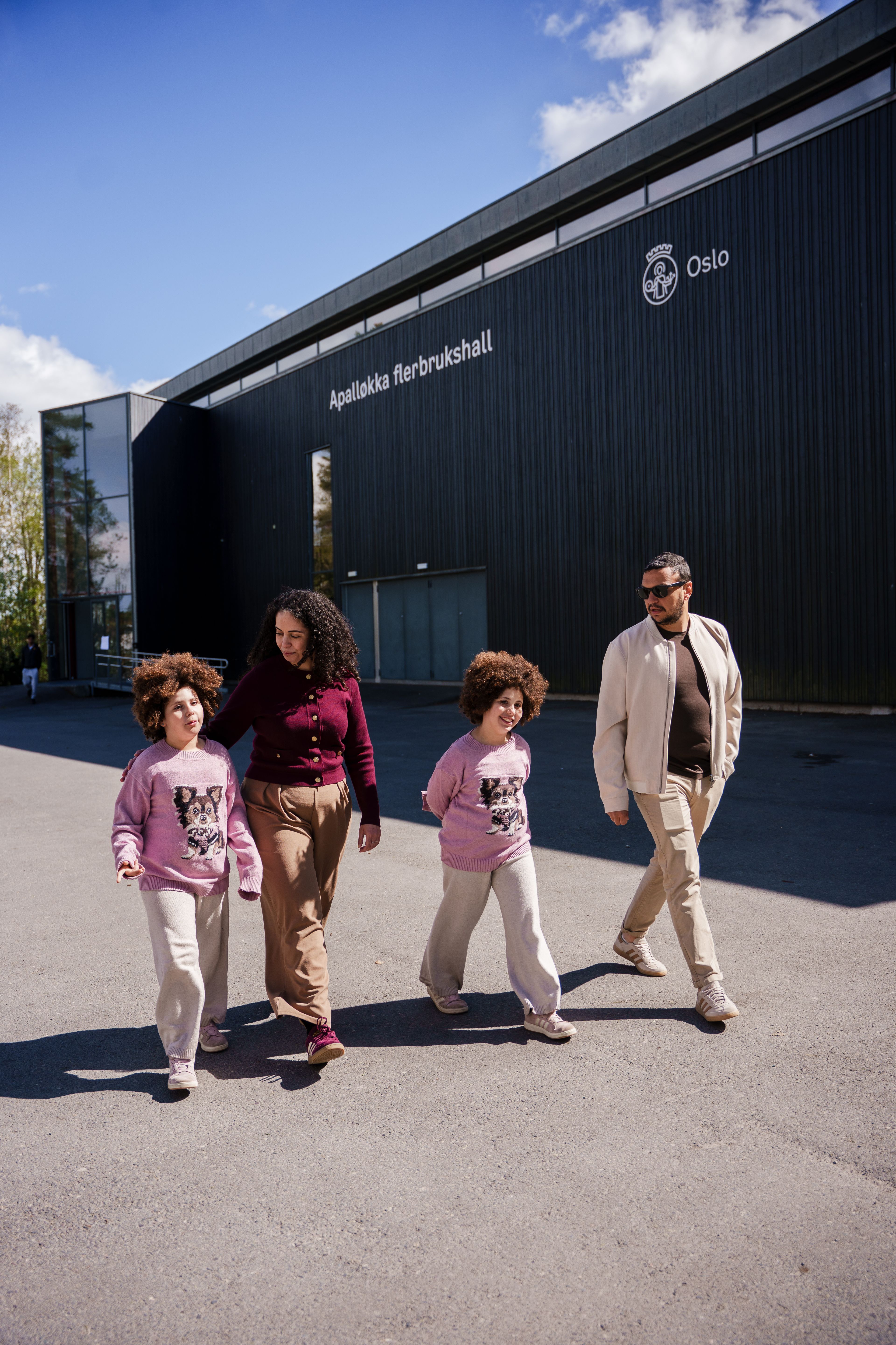 En familie på fire går foran Apalløkka flerbrukshall, en stor, mørk bygning med Oslo kommunes logo på fasaden.