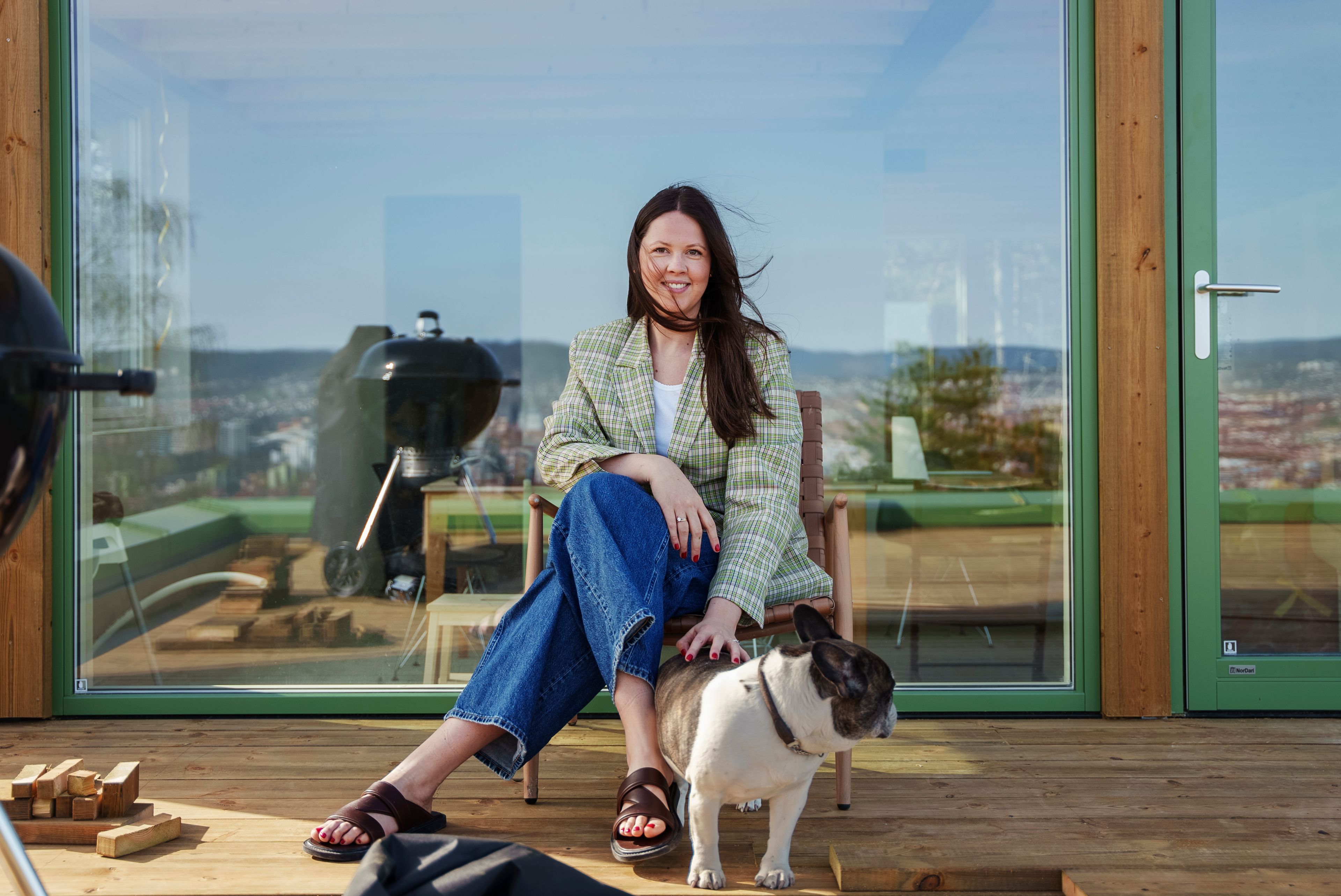 En dame sitter på en lenestol på terrassen. Hun smiler. Håret blåser med vinden. Hun klapper en hund. Det er et stort vindu bak henne med grønne lister.
