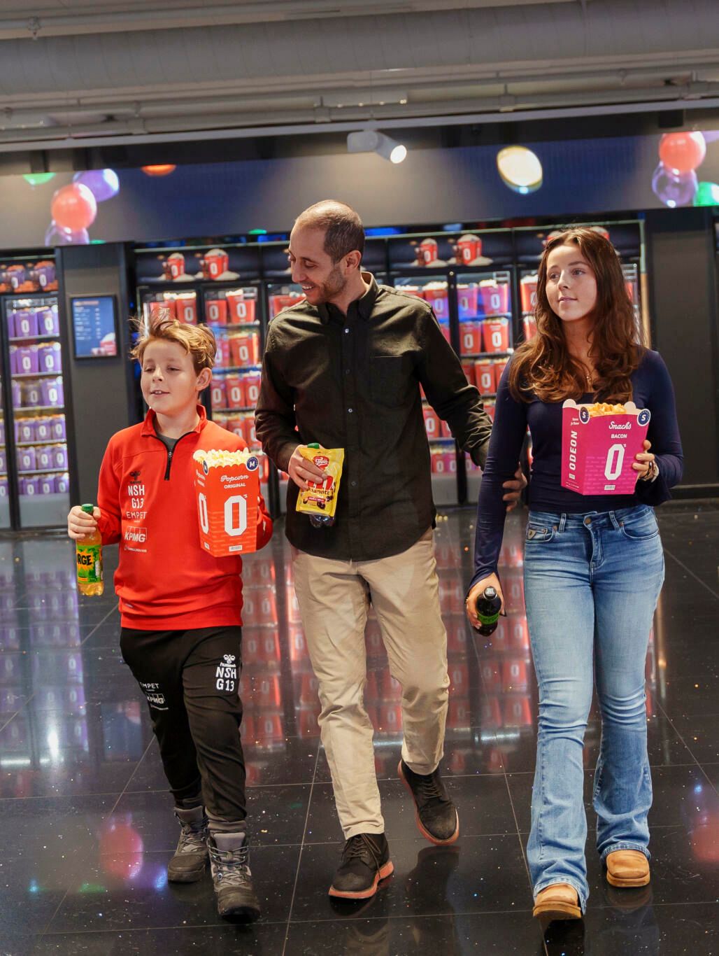 En mann går i midten av to barn og smiler. De er på kino, og barna holder hver sin popcorn og brus.