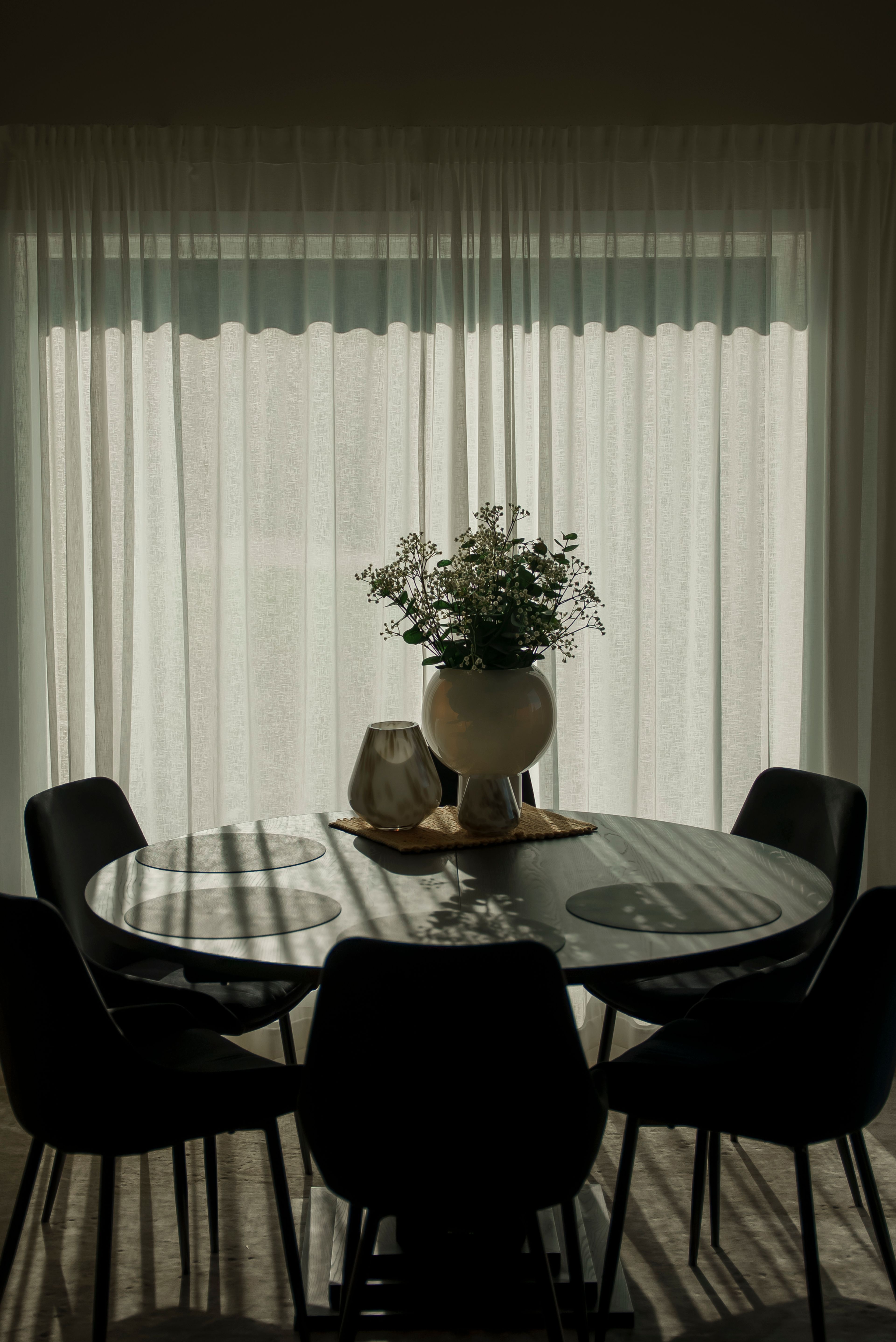 Et mørkt, rundt spisebord foran et vindu tildekt av noen grå gardiner