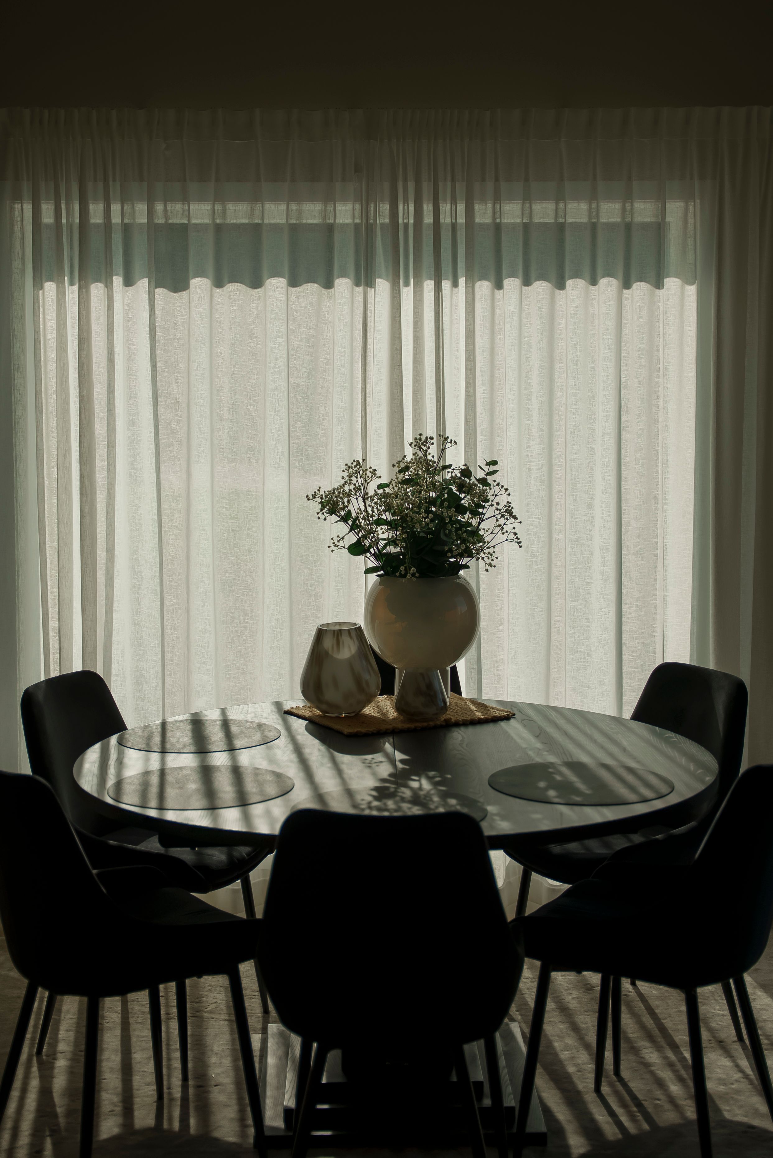 Et mørkt, rundt spisebord foran et vindu tildekt av noen grå gardiner