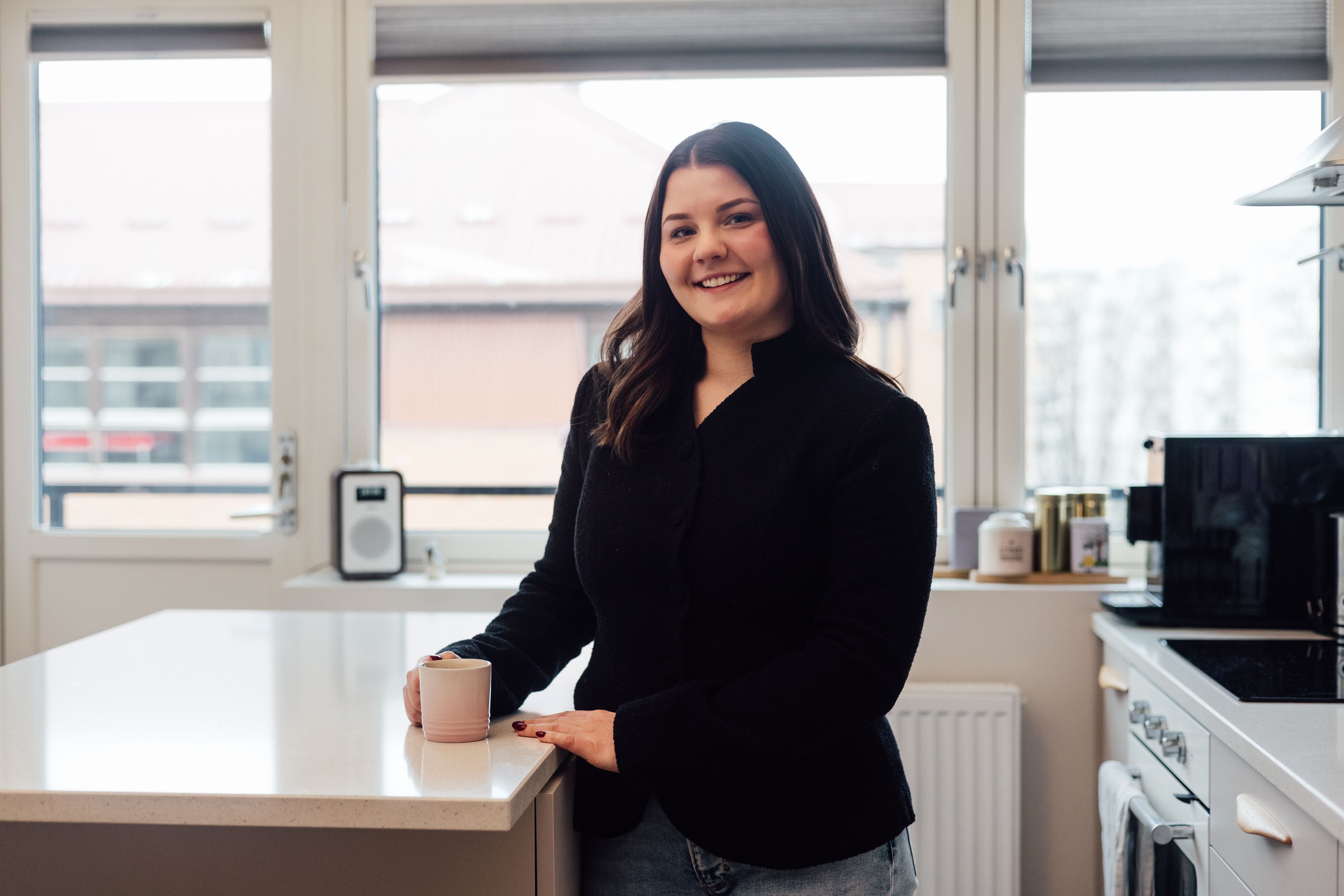 Ei ung dame smiler til kameraet mens hun drikker kaffe ved kjøkkenbenken.