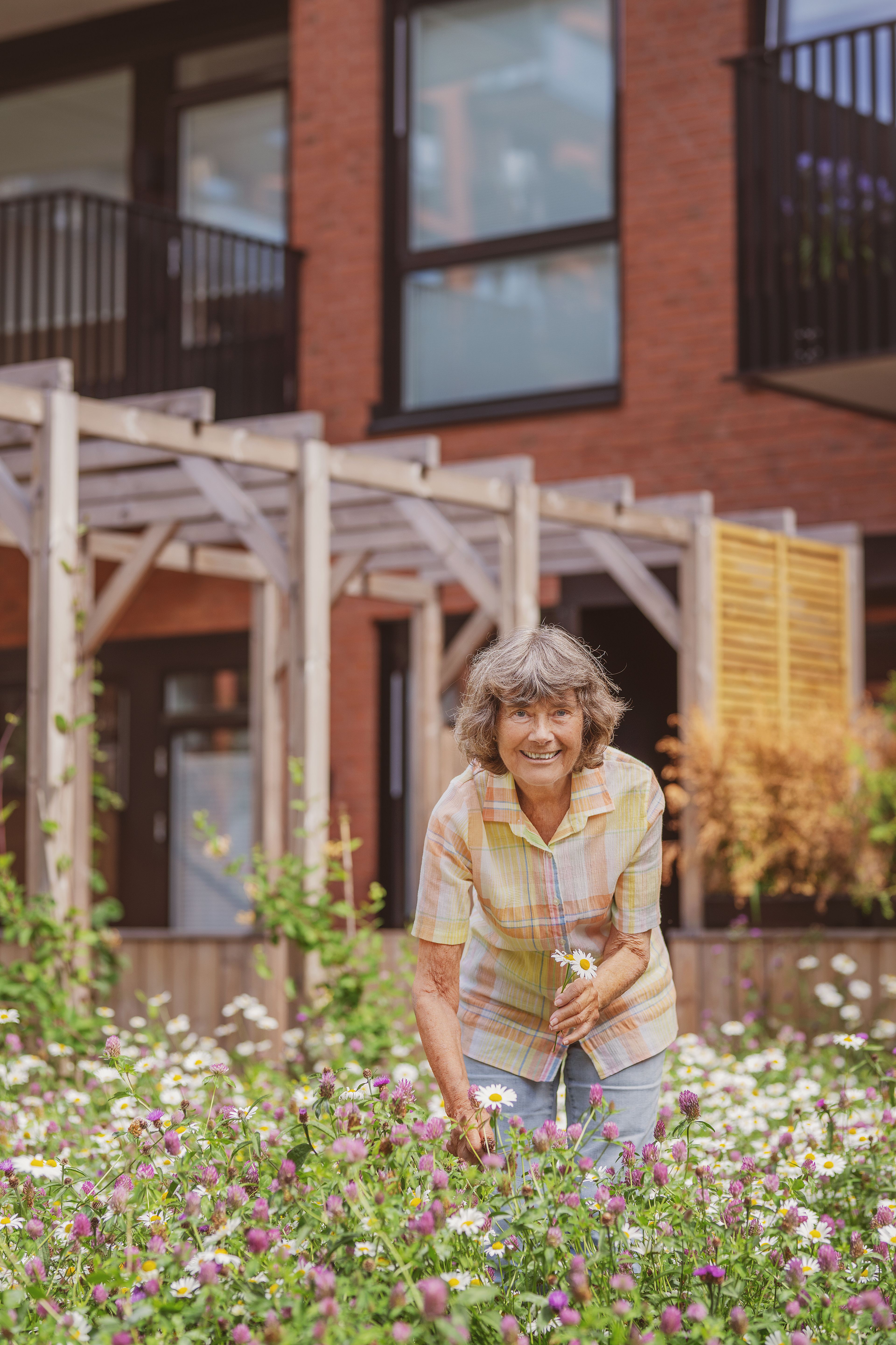 En eldre kvinne smiler til kamera mens hun plukker blomster utenfor et boligbygg.