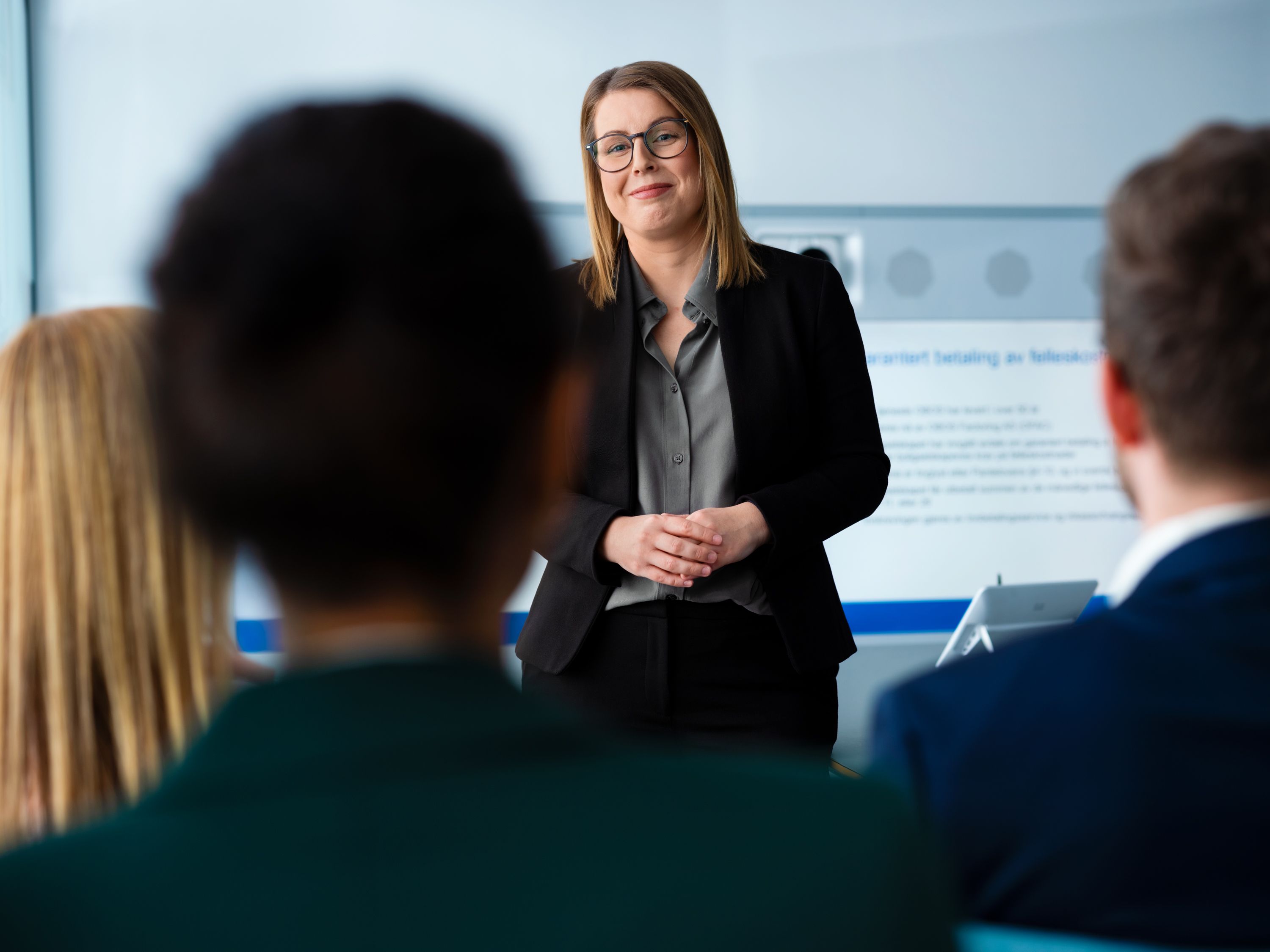 En kvinne i en svart blazer smiler selvsikkert mens hun snakker med tre sittende personer i et lyst rom, noe som antyder et forretningsmøte eller en presentasjonssetting.
