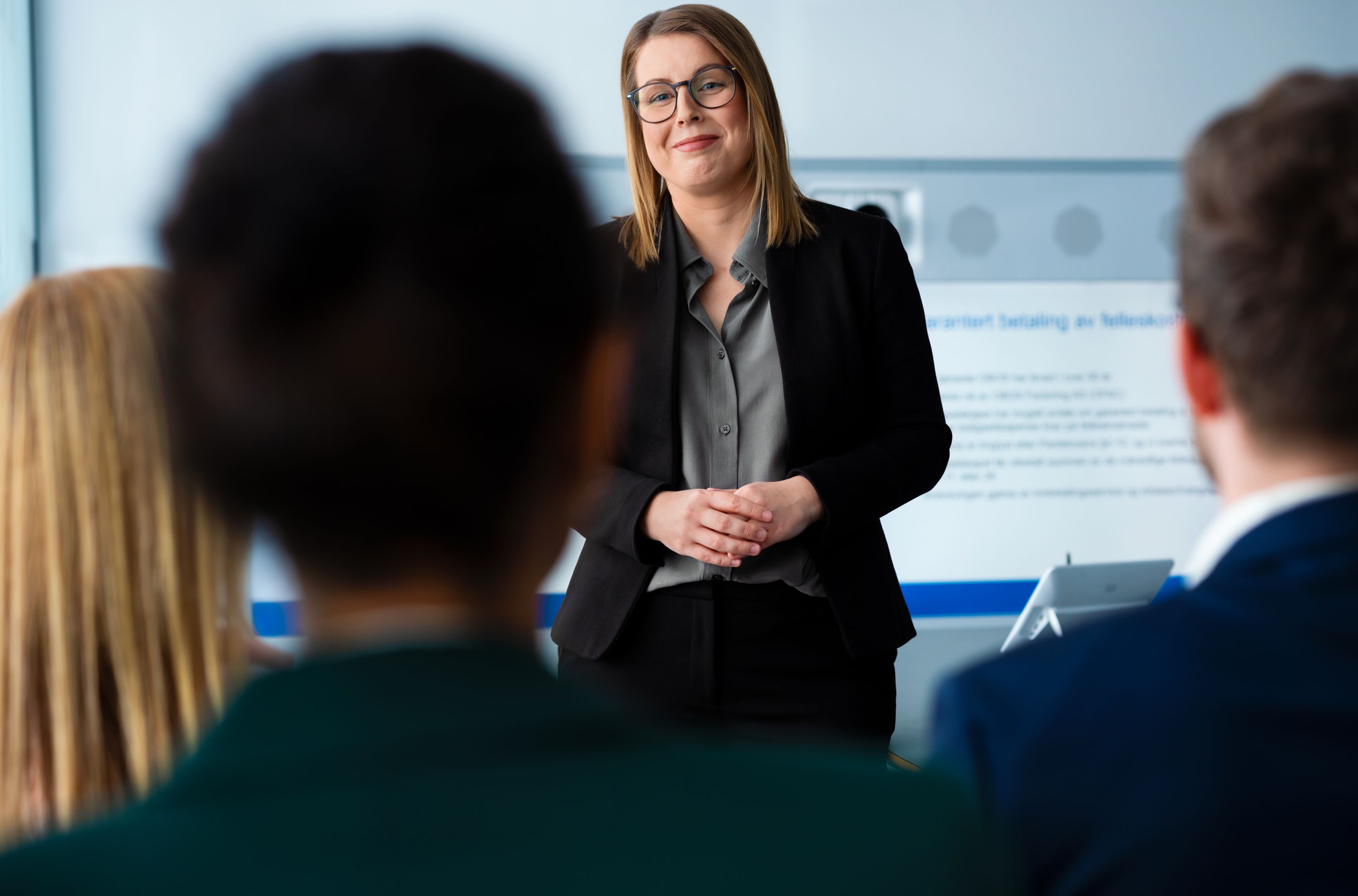 En kvinne i en svart blazer smiler selvsikkert mens hun snakker med tre sittende personer i et lyst rom, noe som antyder et forretningsmøte eller en presentasjonssetting.