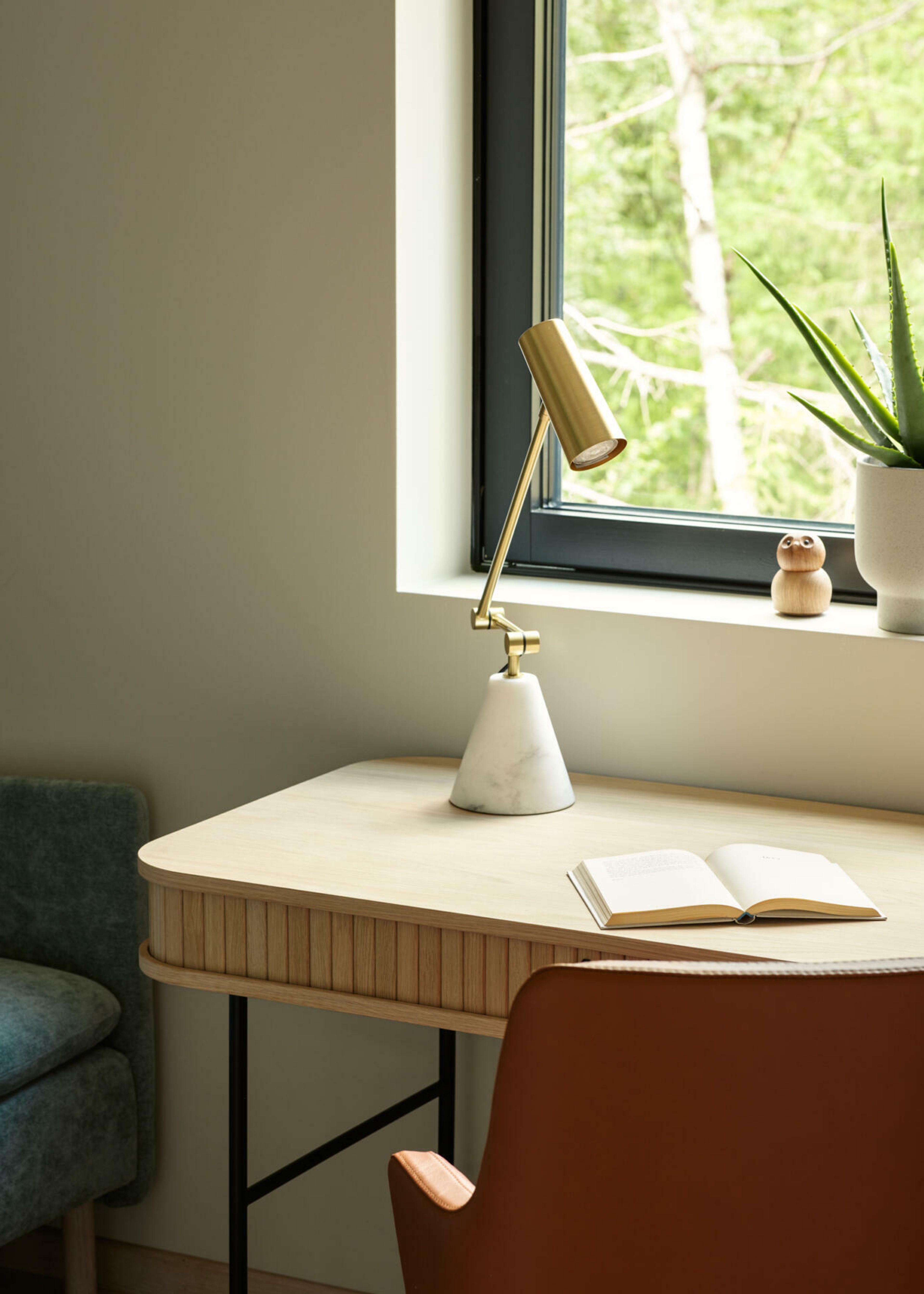 Skrivebord i eik, stol i skinn og en grå sofa på et kontor med utsikt ut over skogen