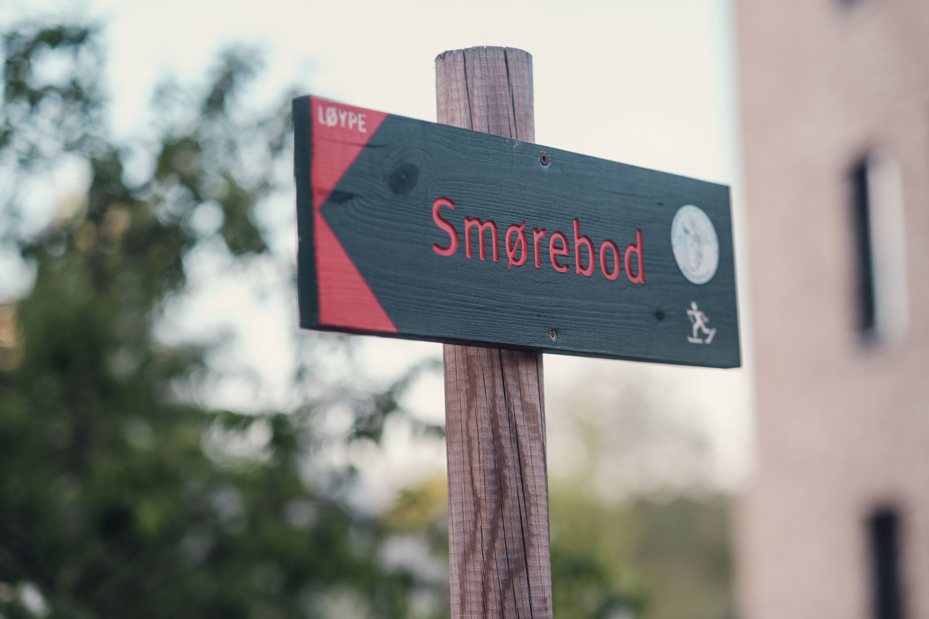 Skiforeningens røde og grønne turskilt med tekst "Smørebod" montert på en trestokk.