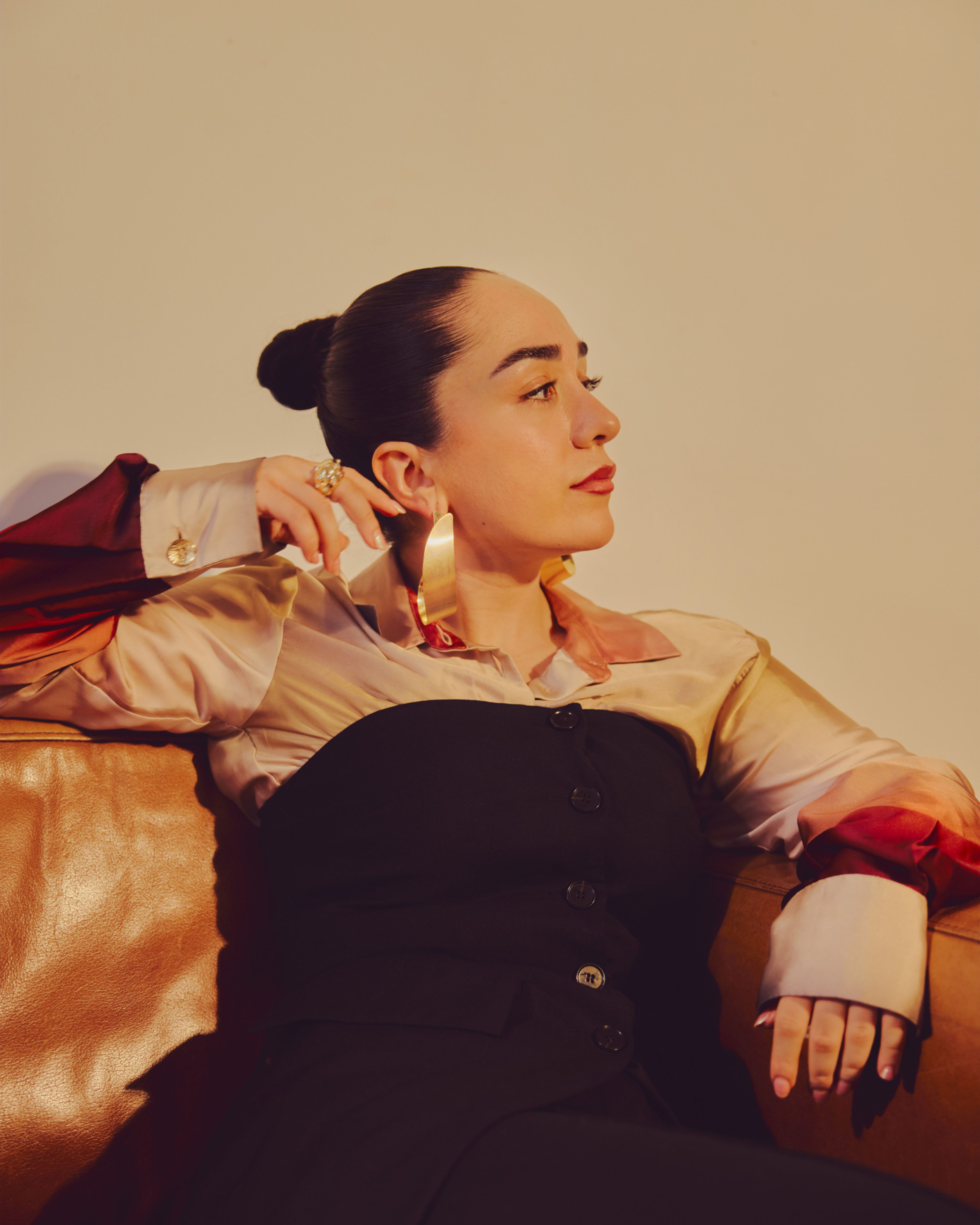 Amanda Delara sitter i en brun skinnsofa. Hun har på seg en silkeskjorte i gull, med en svart kjole over. Hun har store gulløredobber i ørene og håret satt opp i en stram, elegant knute.