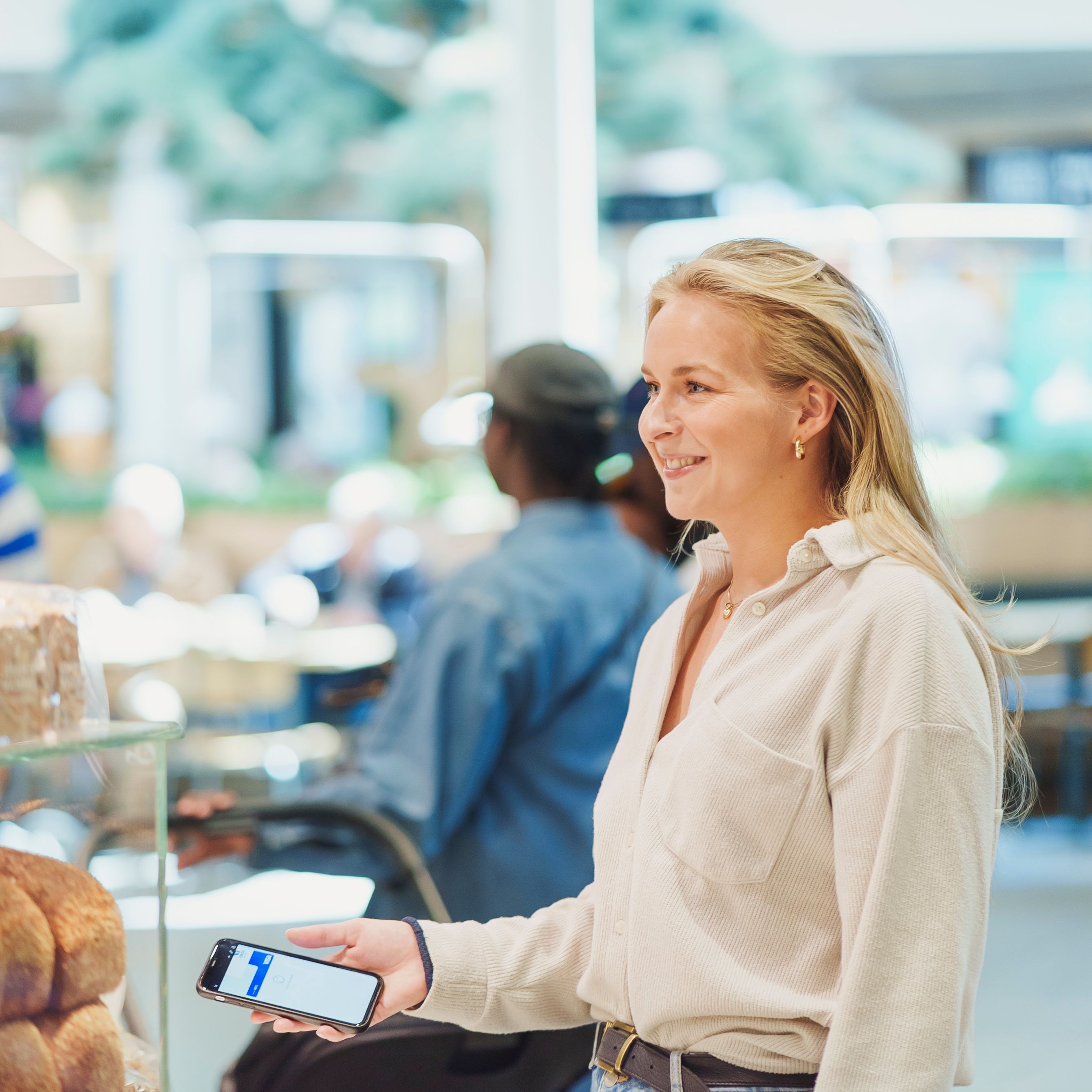 En fornøyd, blond kvinne står i kassen på et bakeri og gjør seg klar for kontaktløs mobilbetaling med Apple Pay