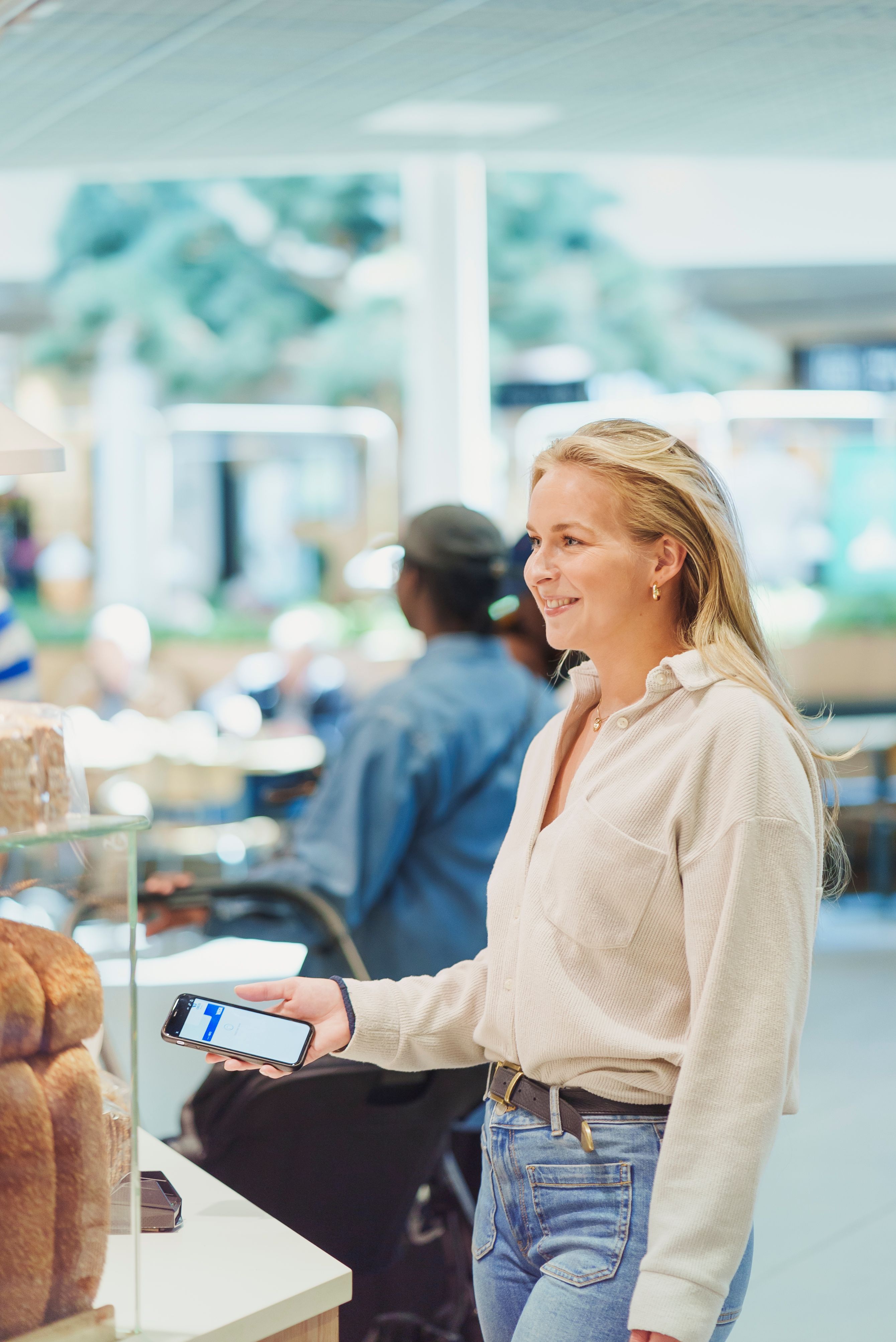 En fornøyd, blond kvinne står i kassen på et bakeri og gjør seg klar for kontaktløs mobilbetaling med Apple Pay